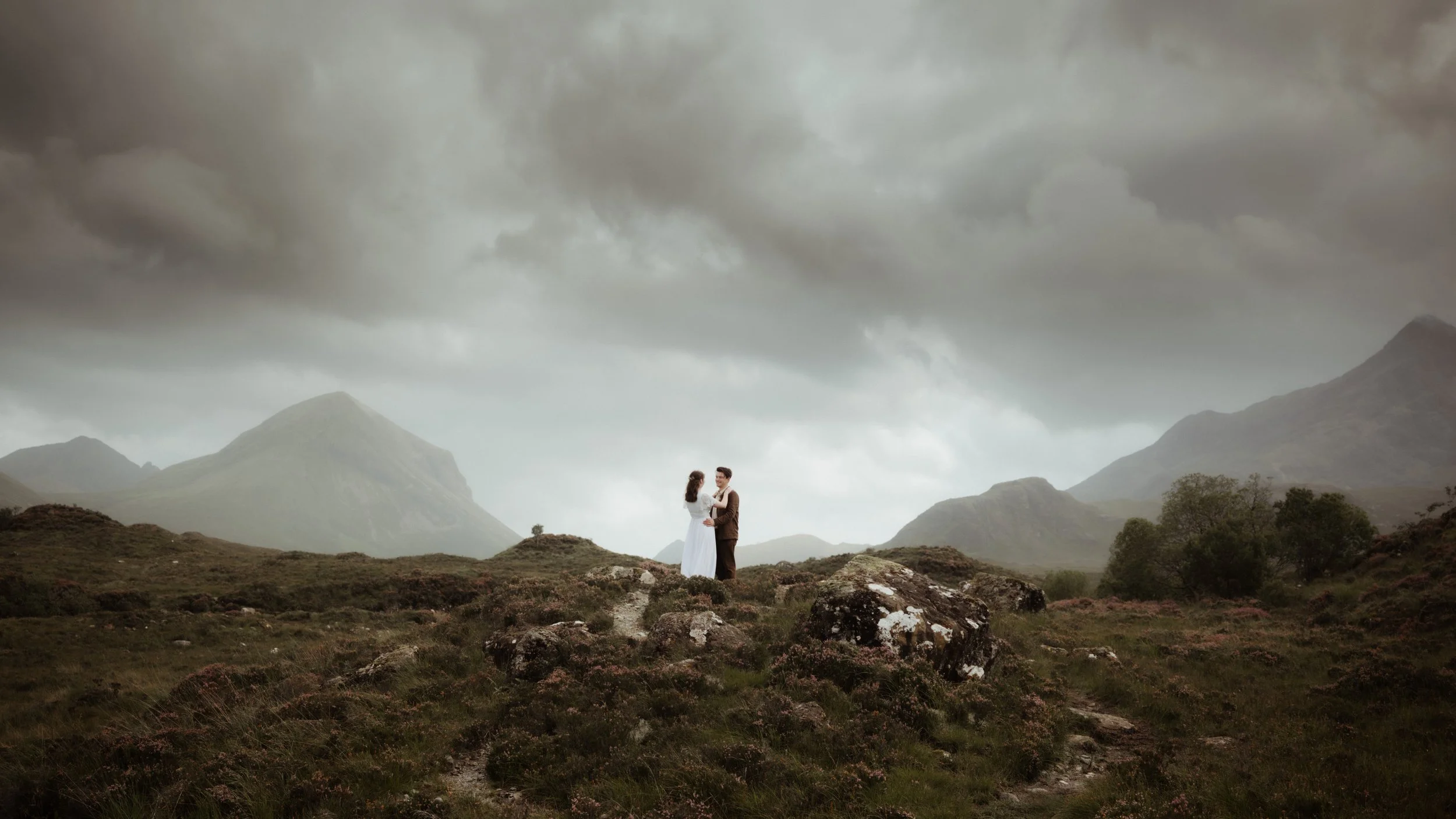 A couple dressed in vintage-style clothing stands on a rocky hillside amid a mountainous landscape with dark, brooding clouds overhead, creating a romantic and dramatic scene.