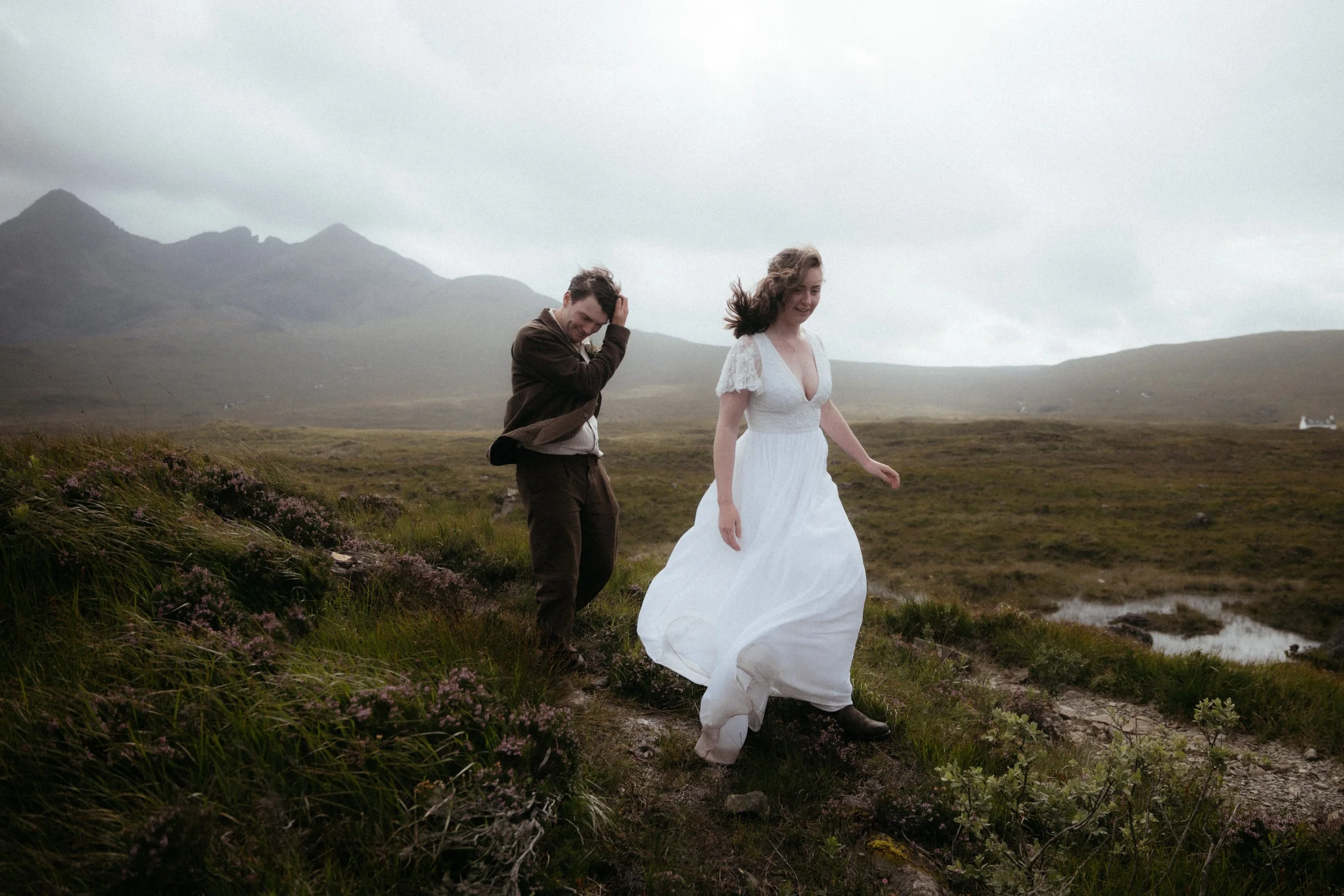 A woman in a white dress walking along a grassy trail next to a man in dark clothing in a mountainous outdoor landscape on a cloudy day.