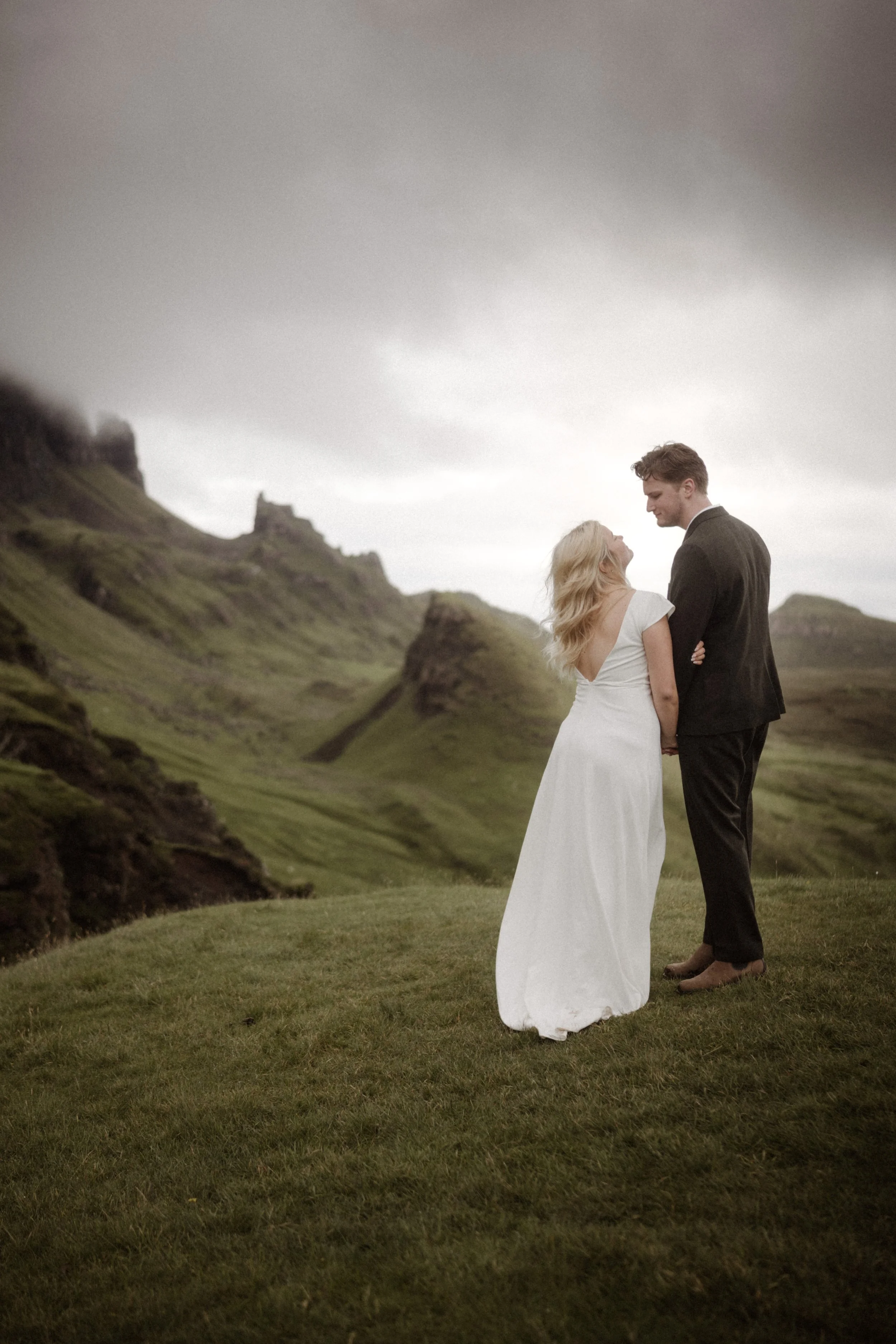 A couple in wedding attire holding hands on a grassy hill with a mountainous landscape in the background.