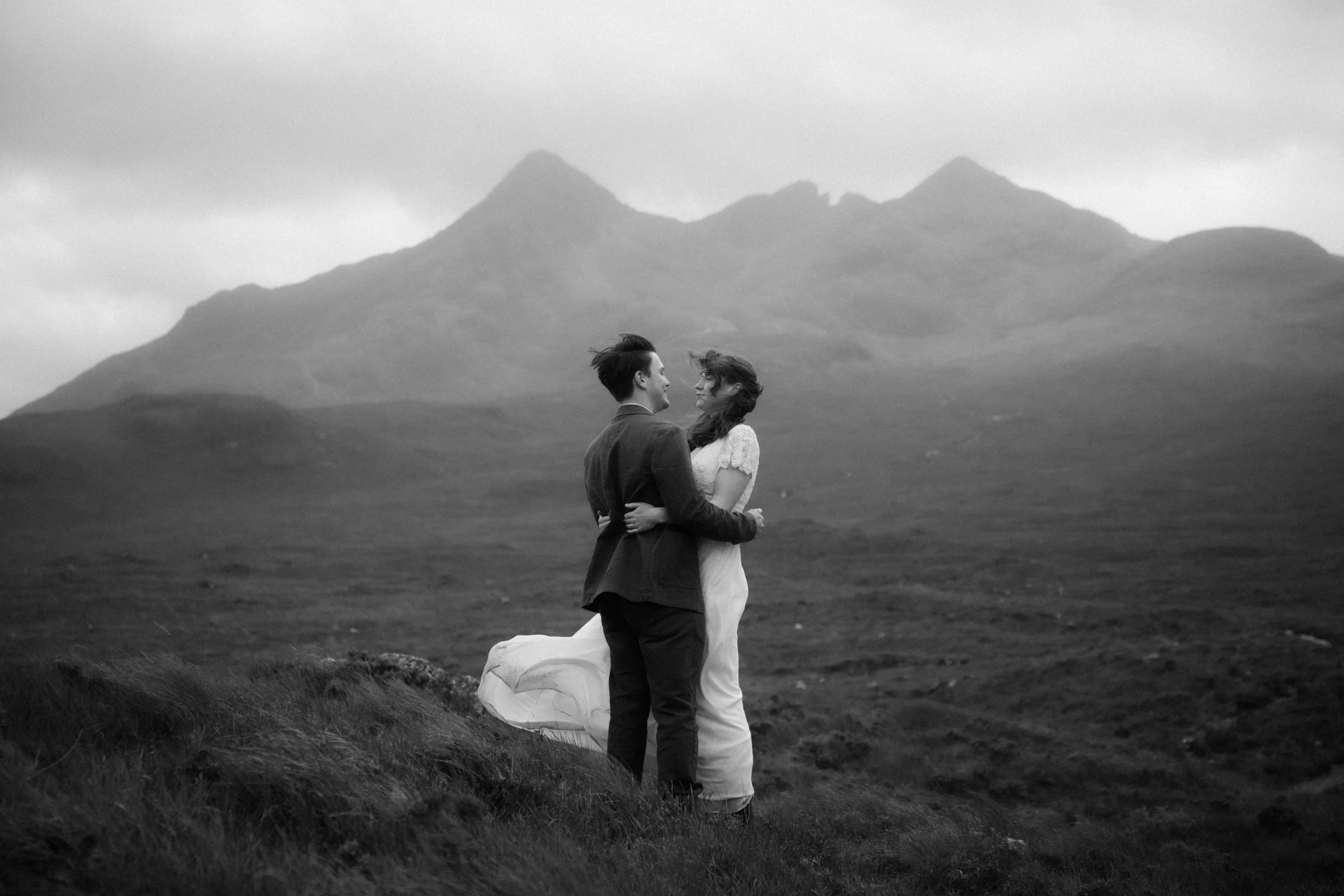 A couple dressed in wedding attire standing close together in a scenic outdoor landscape with mountains in the background, black and white photo.