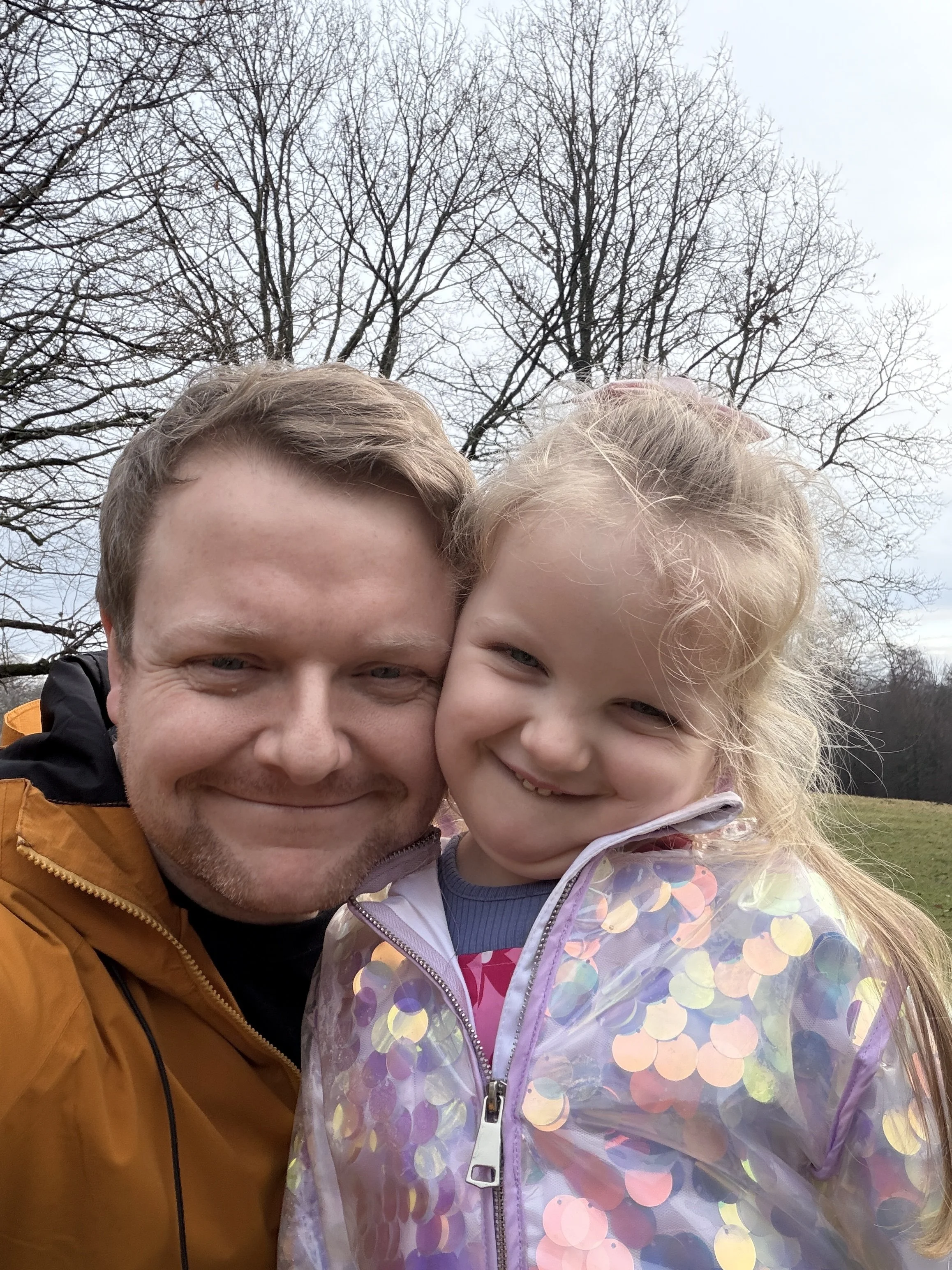 A man and a young girl smiling closely outdoors, with leafless trees and a cloudy sky in the background.