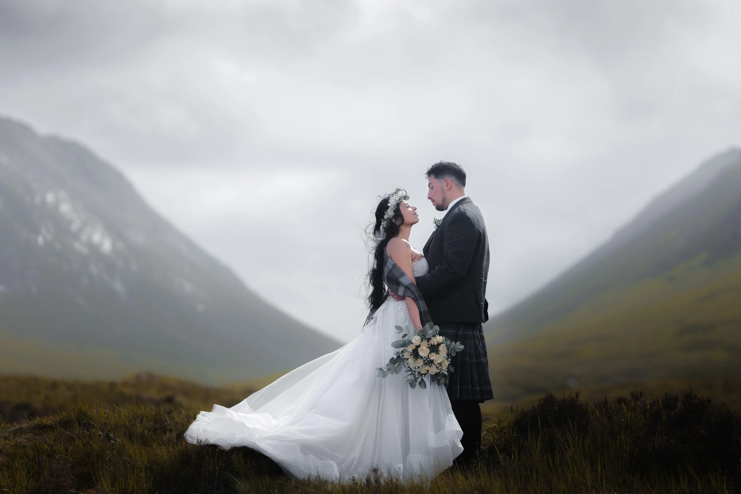A bride and groom standing close together outdoors in a misty mountain landscape, gazing into each other's eyes. The bride is in a white wedding gown holding a bouquet, and the groom is in a dark suit with a kilt.
