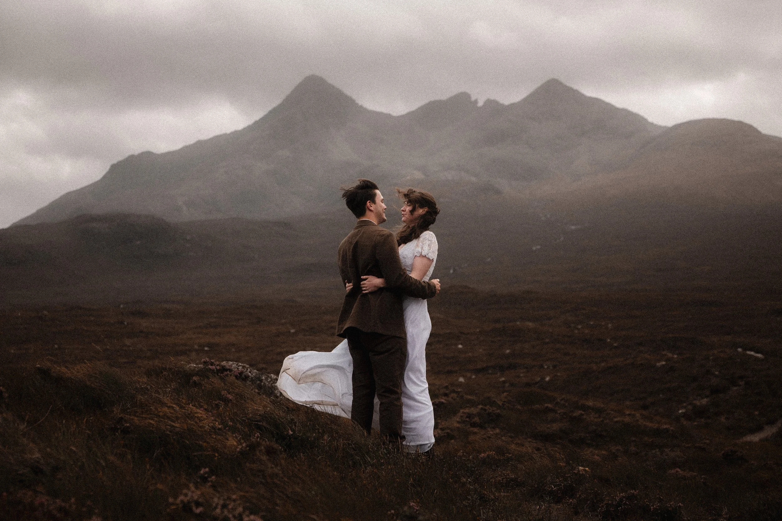 A couple in wedding attire standing on a grassy area in a mountainous landscape on the Isle of Skye with overcast sky, embracing and looking into each other's eyes.