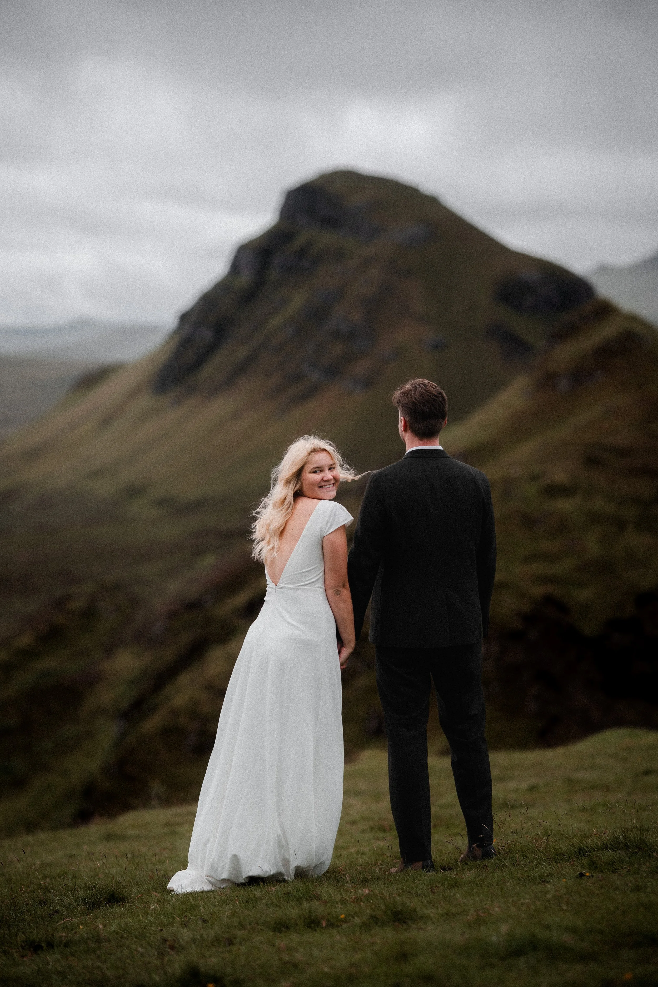 A bride and groom holding hands outdoors on a grassy hill with a mountain in the background on the Isle of Skye.