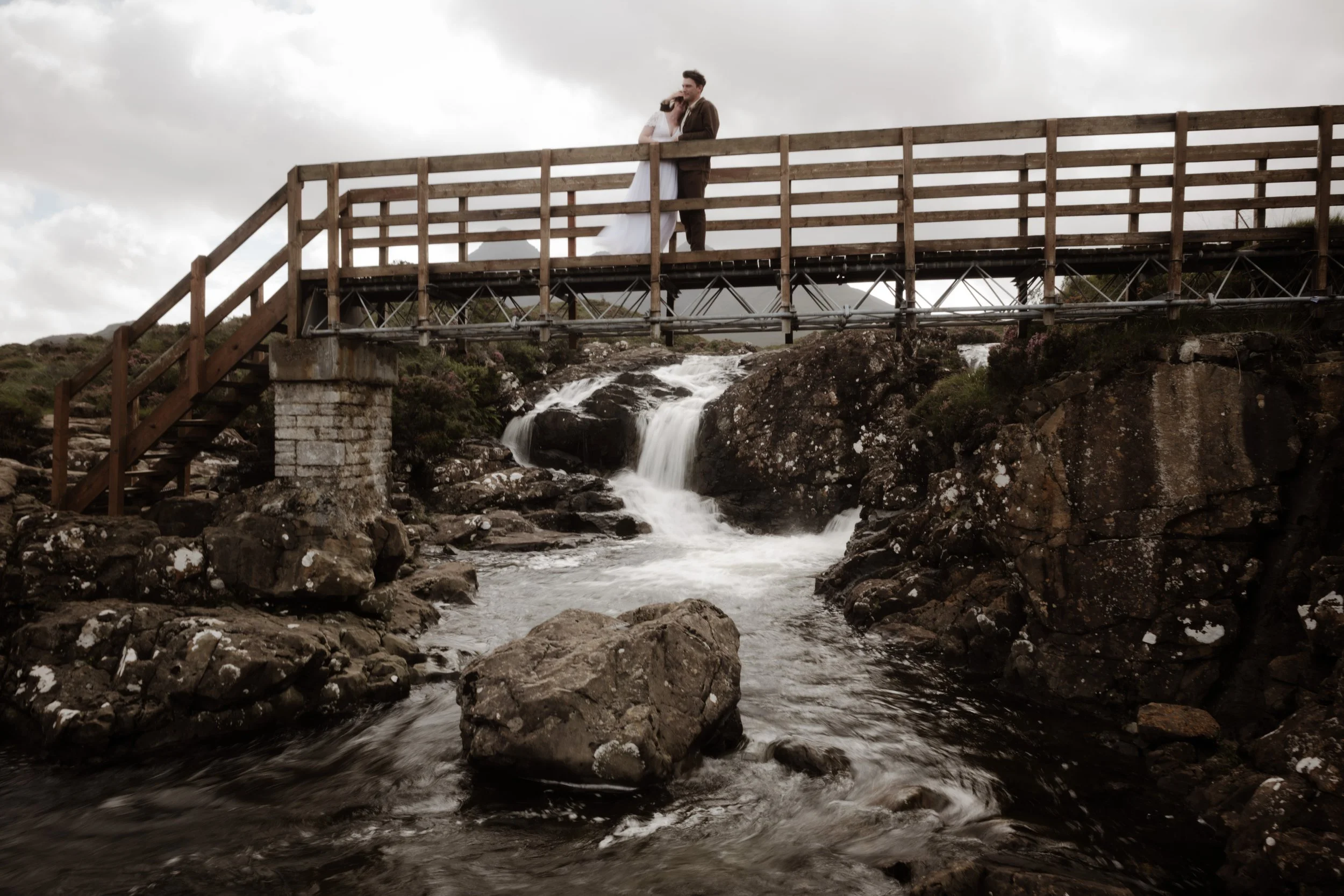A man and woman in wedding attire stand on a wooden bridge over a rocky stream with small waterfalls, under cloudy sky.