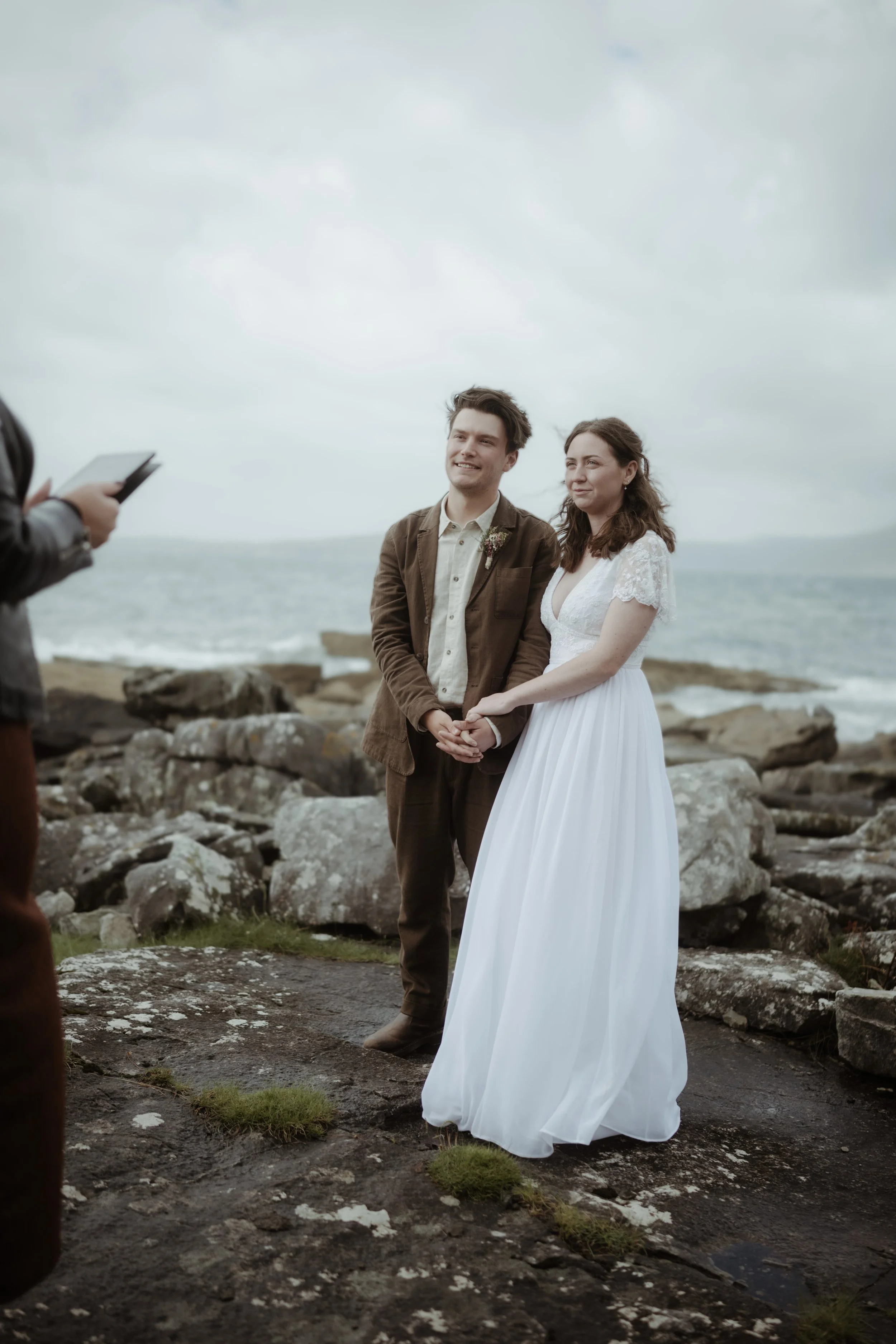 A couple getting married outdoors by the rocks on a cloudy day, the woman in a white wedding dress and the man in a brown suit, holding hands with a person taking their photo.