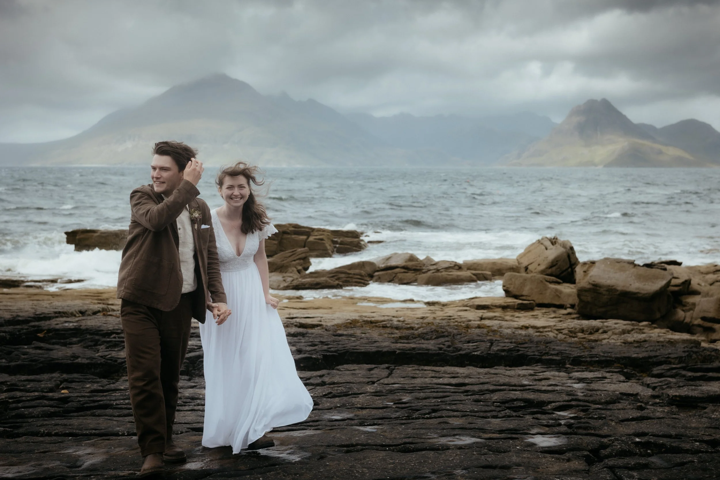 A happy couple walks hand in hand along a rocky beach, with the ocean and mountains in the background on a cloudy day.