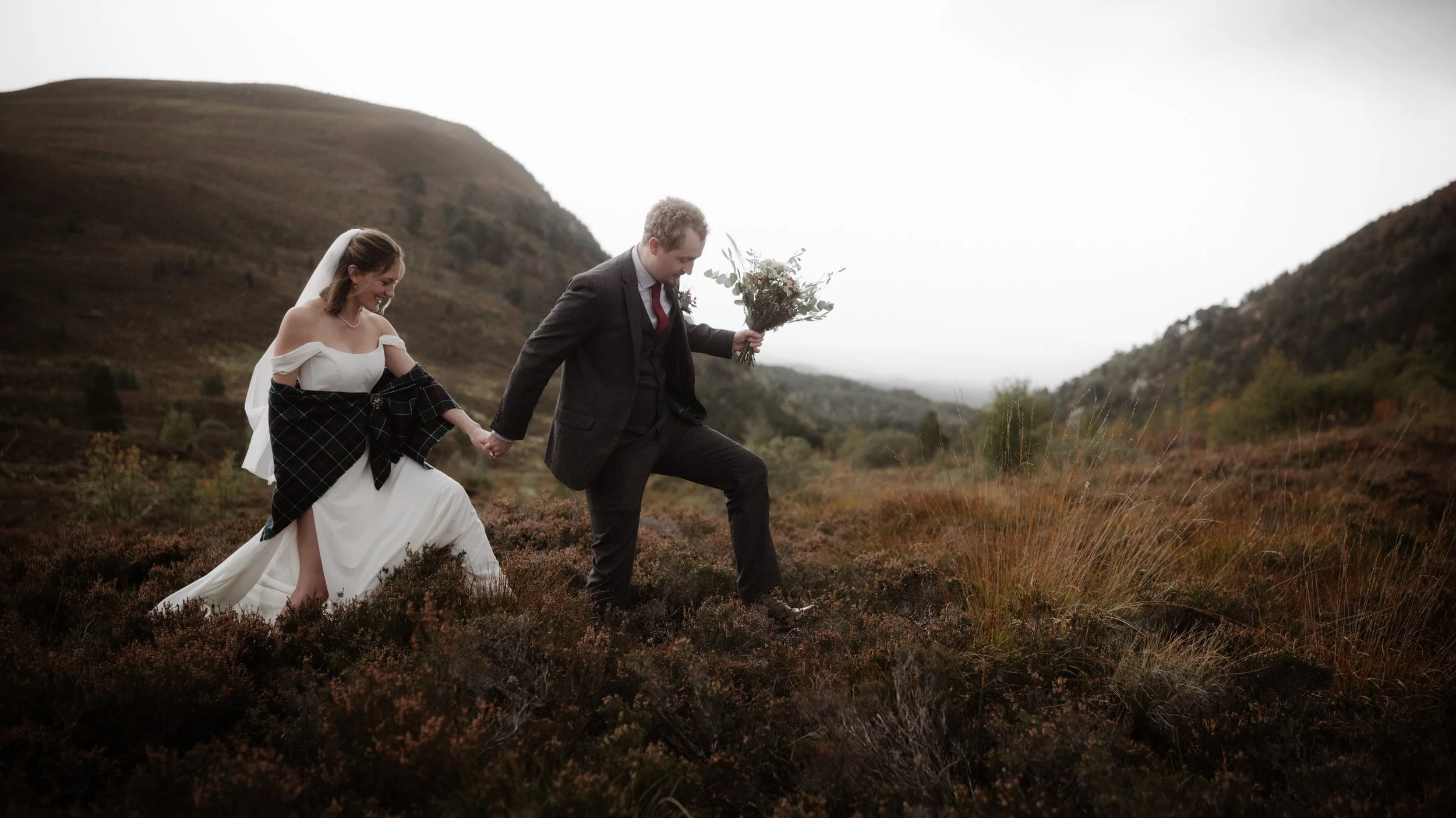A couple dressed in wedding attire walking through a grassy, hilly landscape on a cloudy day, holding hands, with the groom holding a bouquet of flowers.