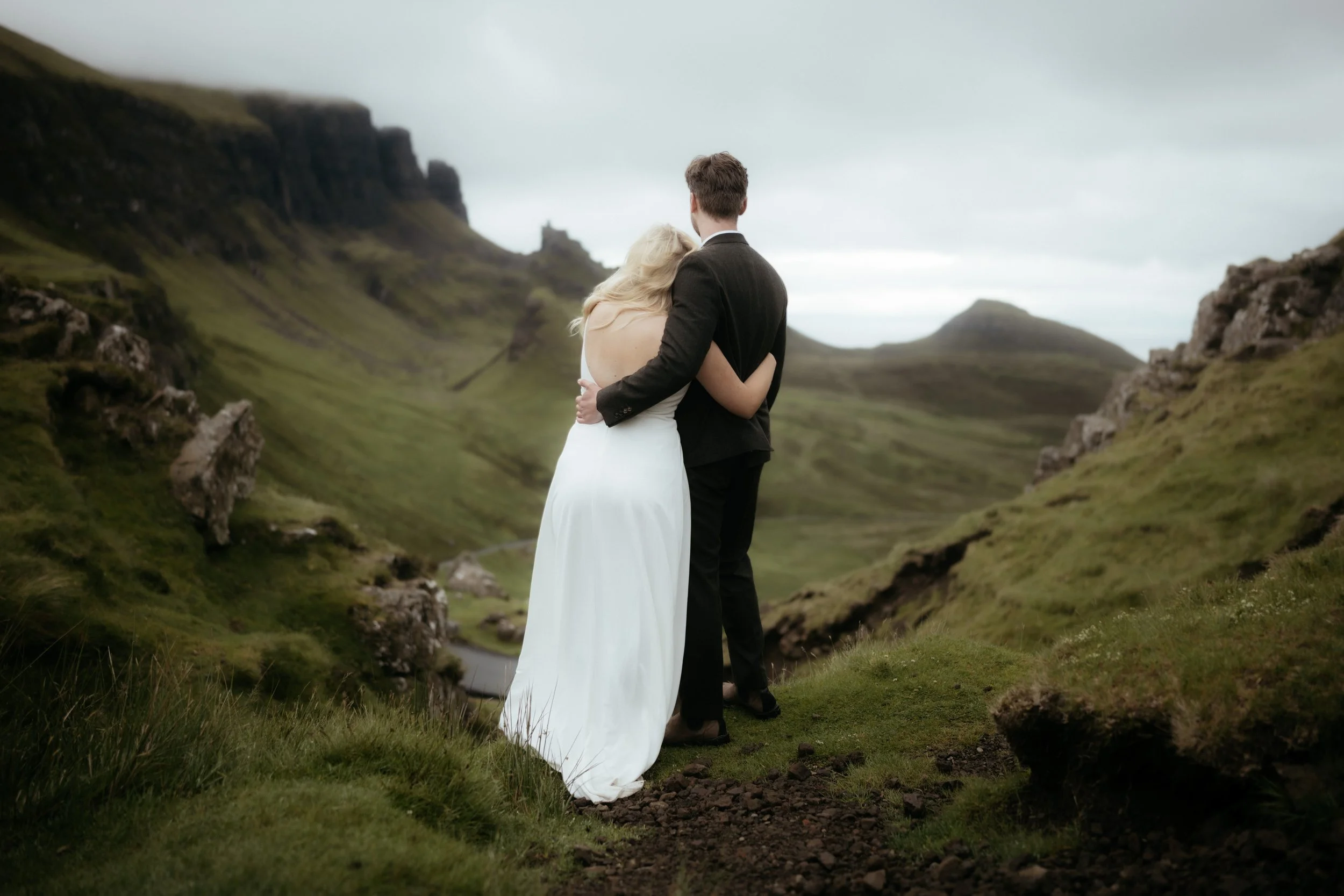 A bride and groom standing together on a grassy hillside, embracing with mountains in the background.