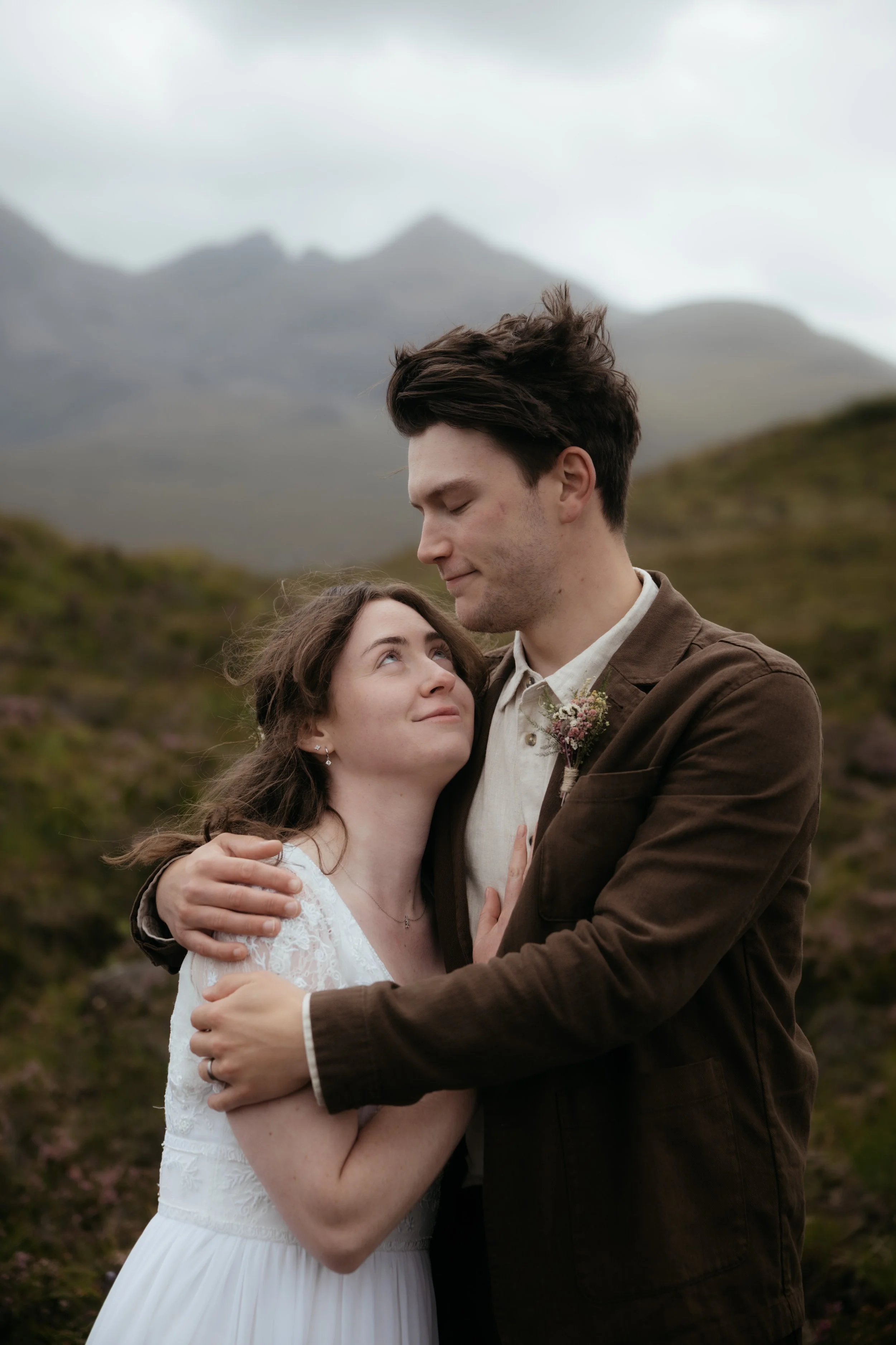 A young couple embracing outdoors in a mountainous area during overcast weather, with the woman gazing affectionately at the man, who is looking down at her.