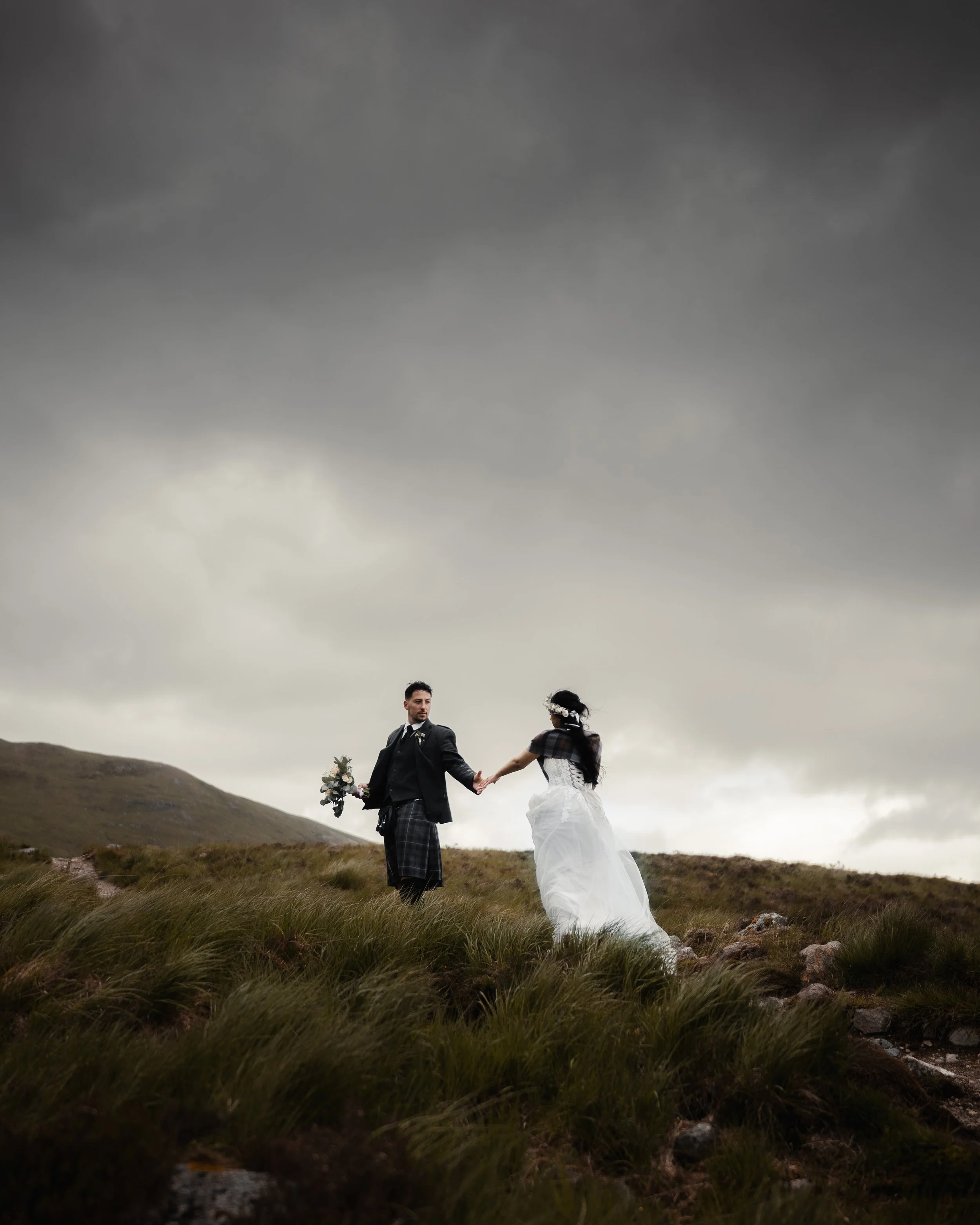 A bride and groom holding hands outdoors on a grassy hill with mountains and dark cloudy sky in the background.