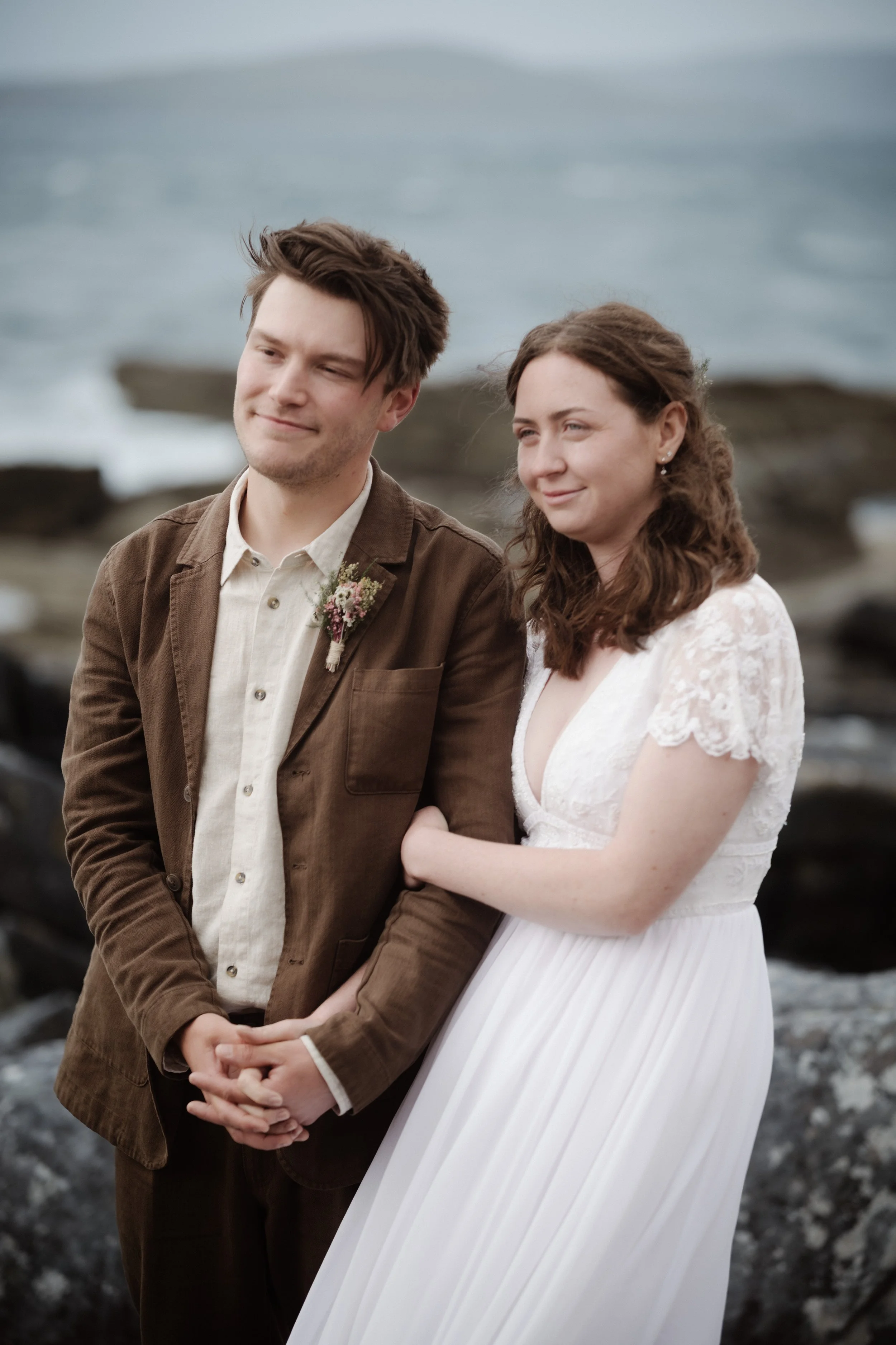 A young couple dressed in wedding attire standing near the rocky shoreline with the ocean in the background. The man is wearing a brown jacket and a boutonniere, while the woman is in a white lace wedding dress with her arm linked around his.