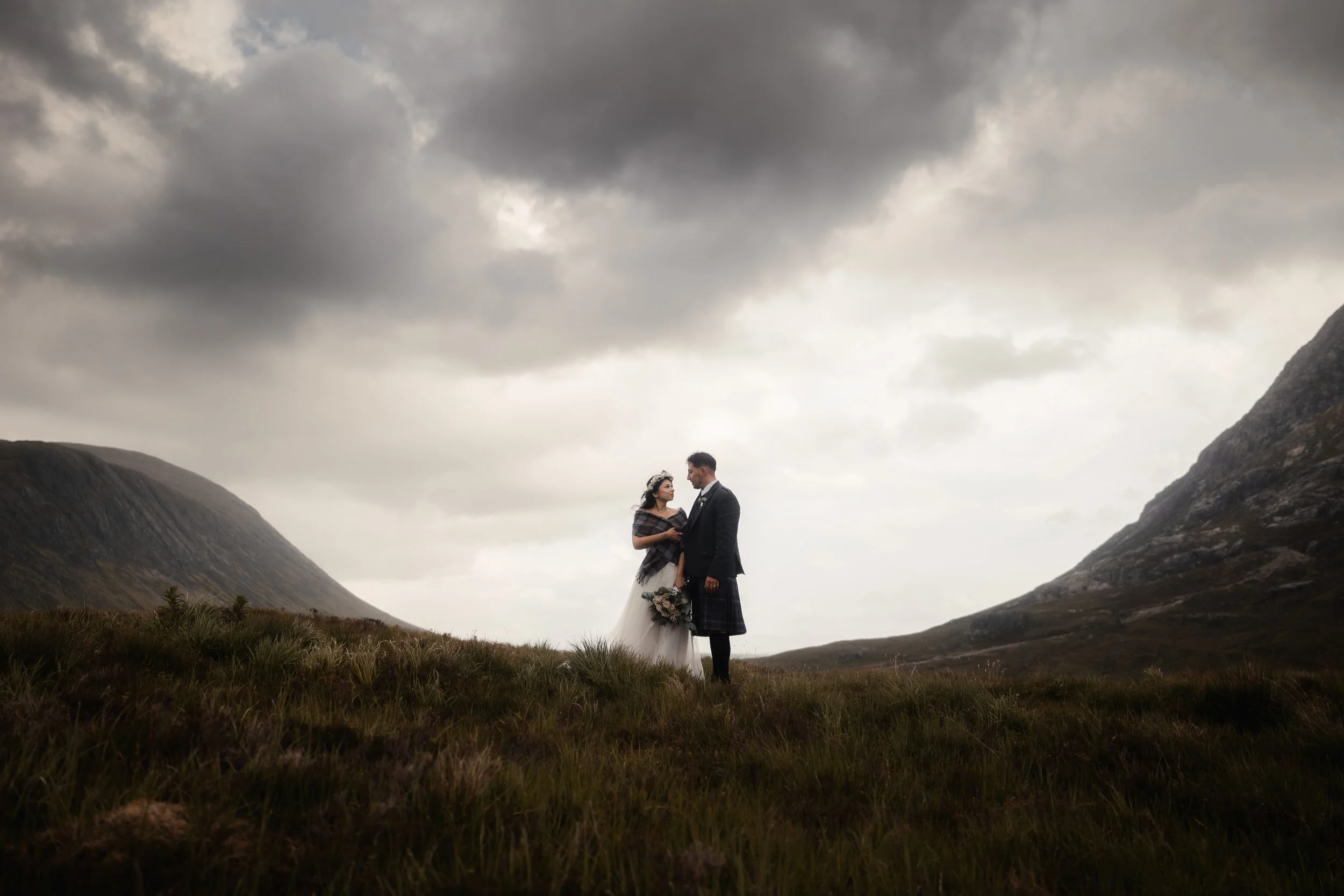 A bride and groom stand together on a grassy hillside with mountains in the background, under a cloudy sky, during a wedding photoshoot.