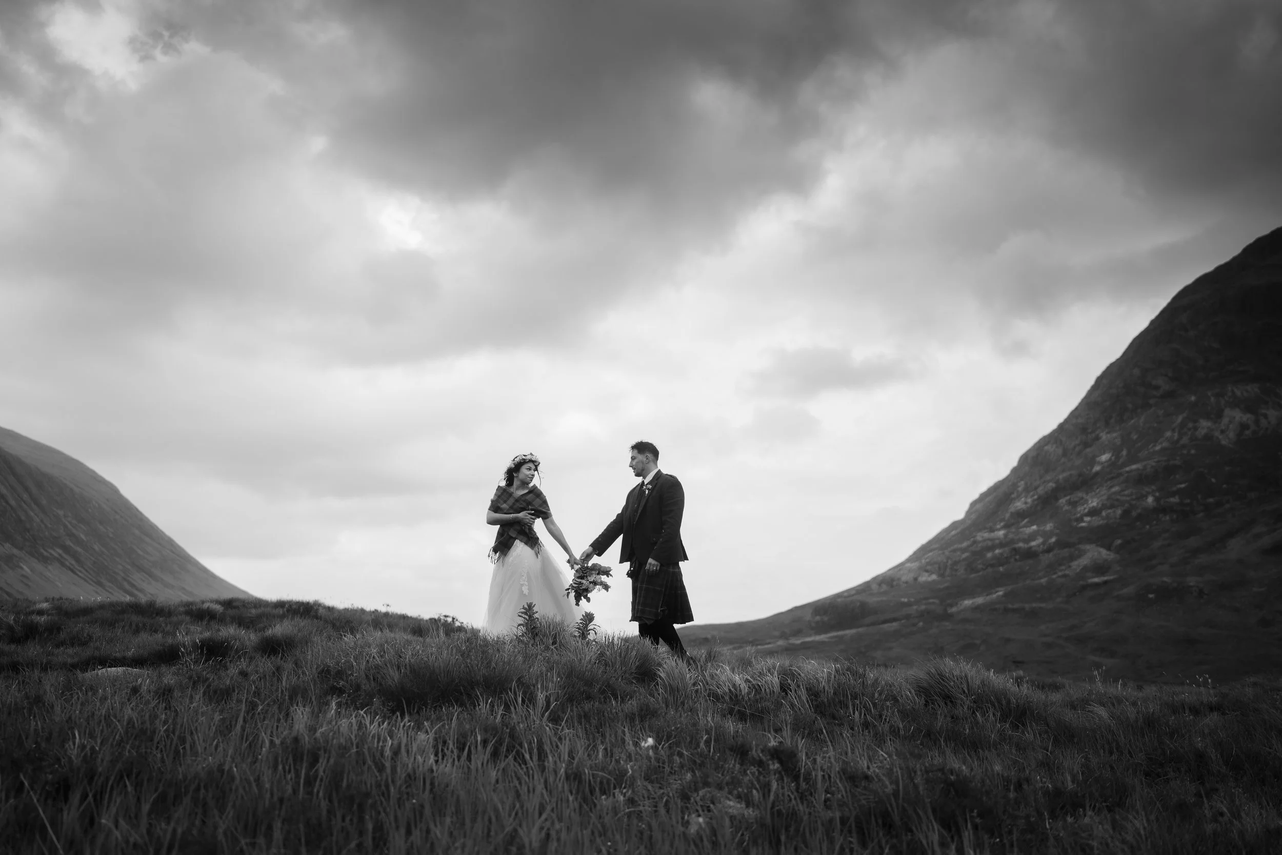 A black and white photo of a couple dressed in wedding attire standing outdoors on a grassy field, holding hands and facing each other with mountains and a cloudy sky in the background.