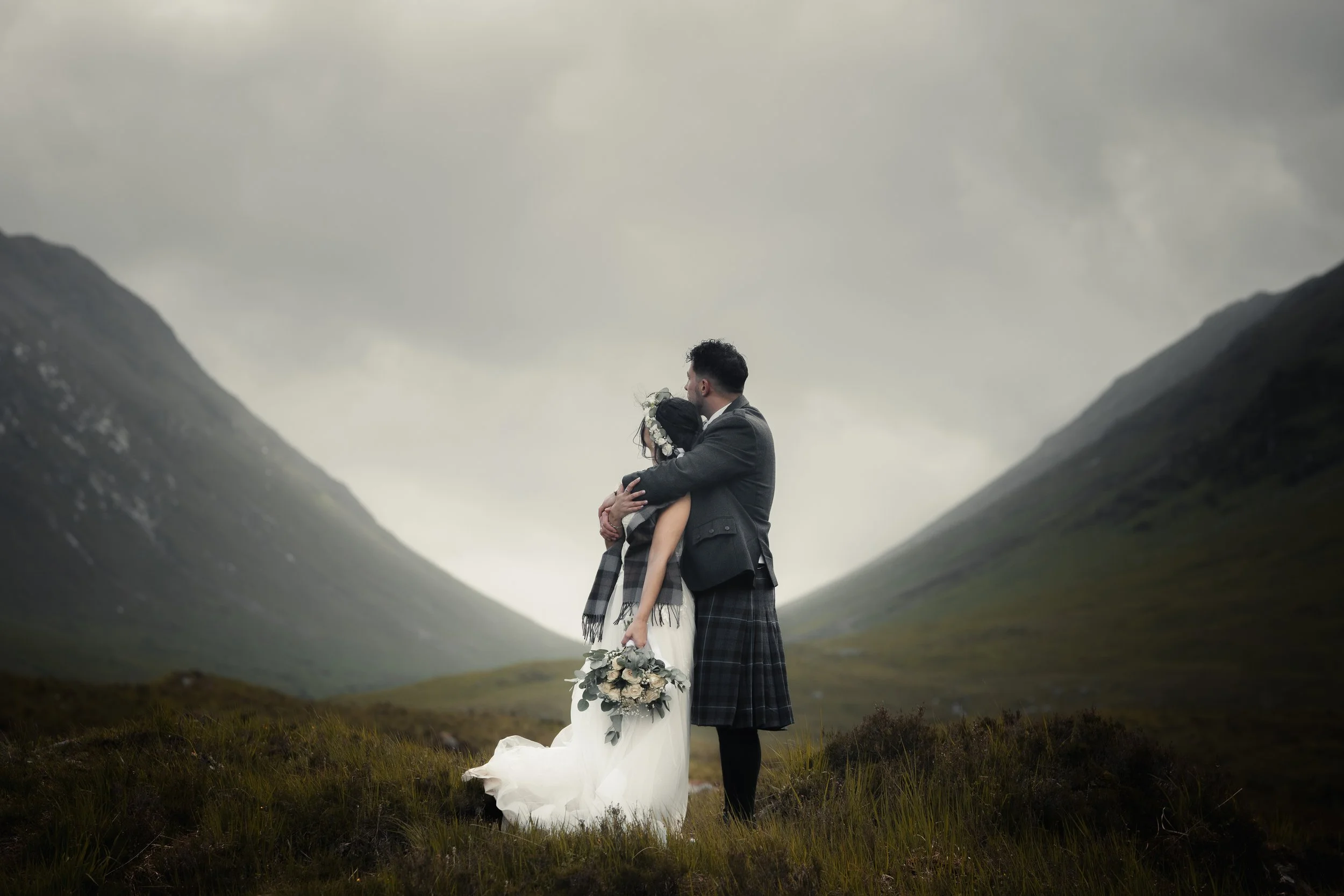A couple dressed in wedding attire embracing in a scenic mountainous landscape under a cloudy sky, with the bride holding a bouquet of flowers.