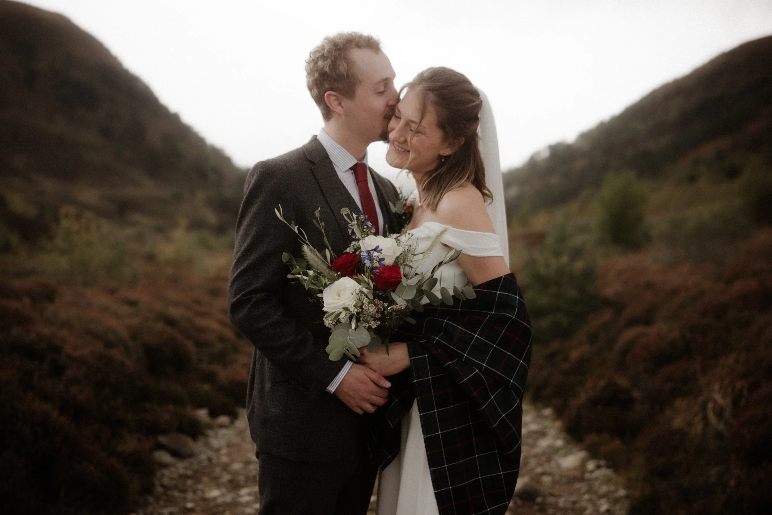 A newlywed couple sharing an intimate moment outdoors, with the groom kissing the bride on the forehead, standing in a natural setting with hills in the background. The bride is holding a bouquet of flowers and is dressed in a white off-shoulder wedd