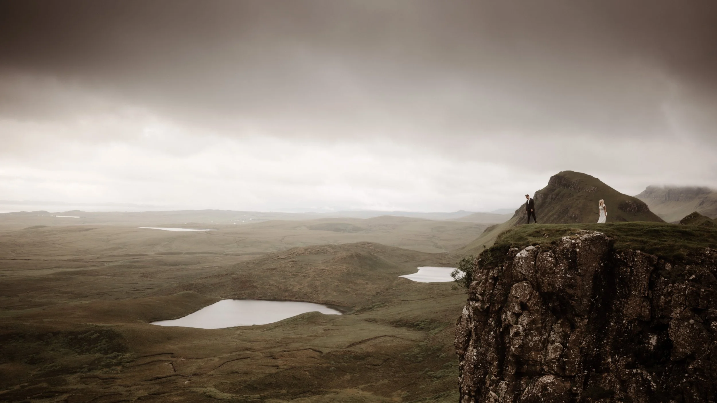 A man and woman standing on a grassy cliff edge overlooking a vast moorland with small lakes, under a cloudy sky.