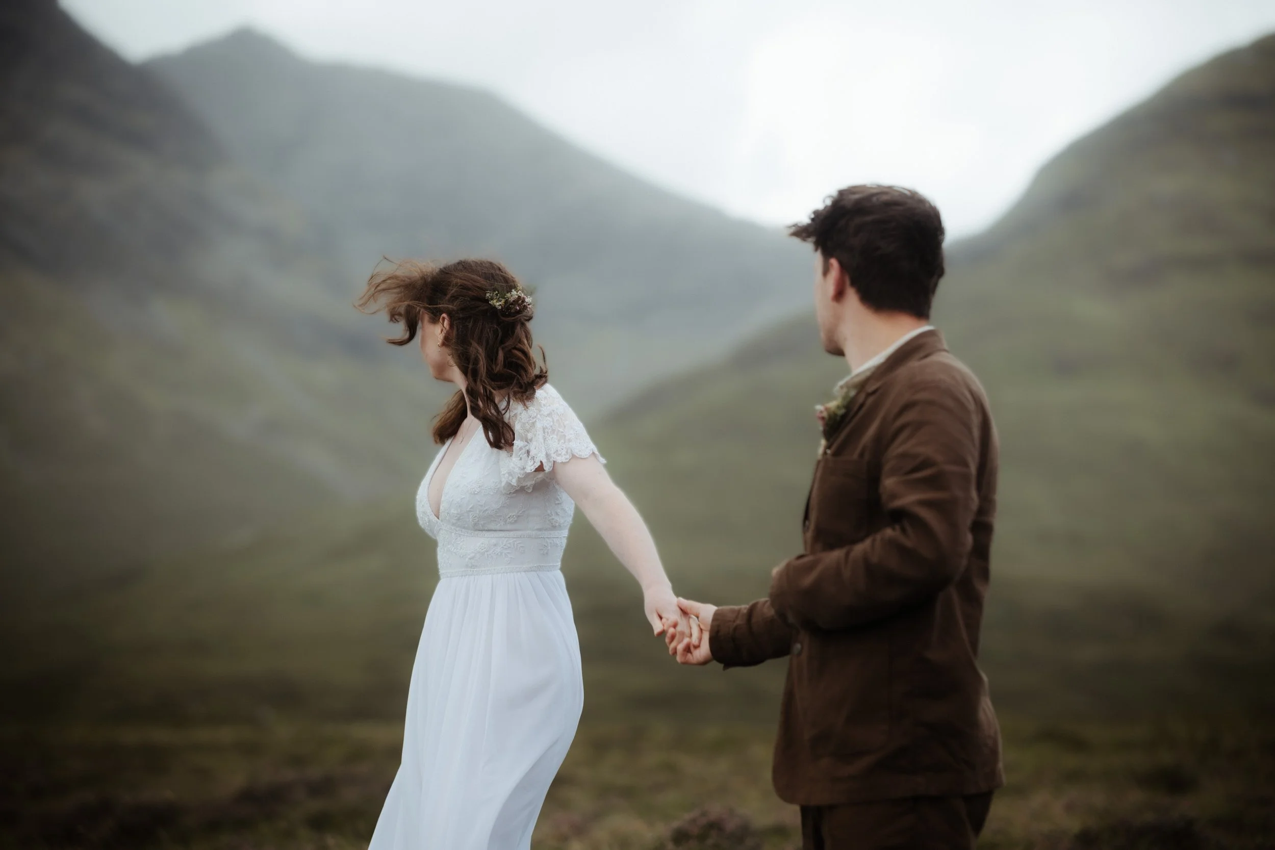 A couple holding hands in a scenic, foggy mountainous landscape, with the woman wearing a white dress and the man in a brown suit.