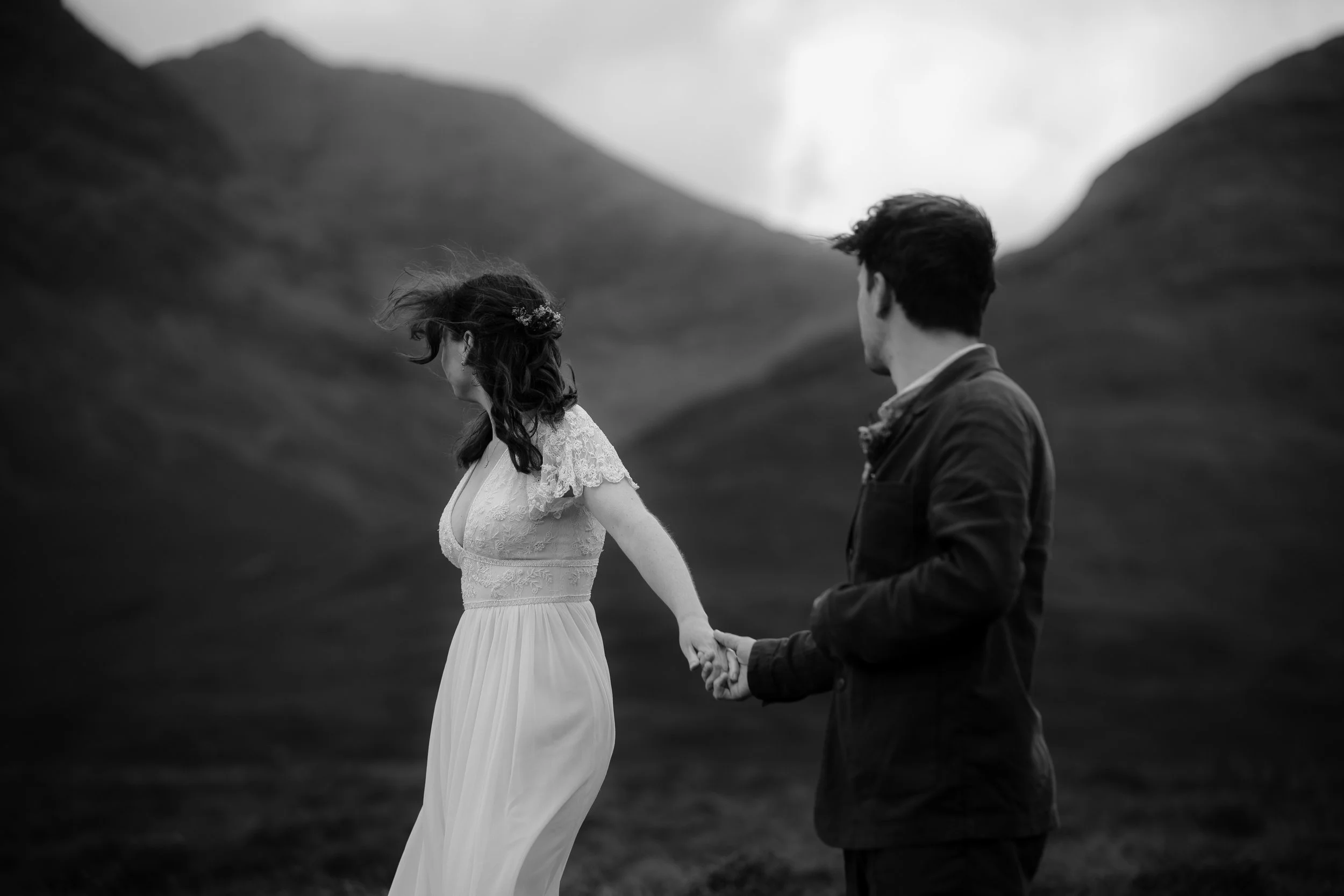 A black and white photo of a couple holding hands on the Isle of Skye, with the woman in a white dress and the man in a dark jacket.