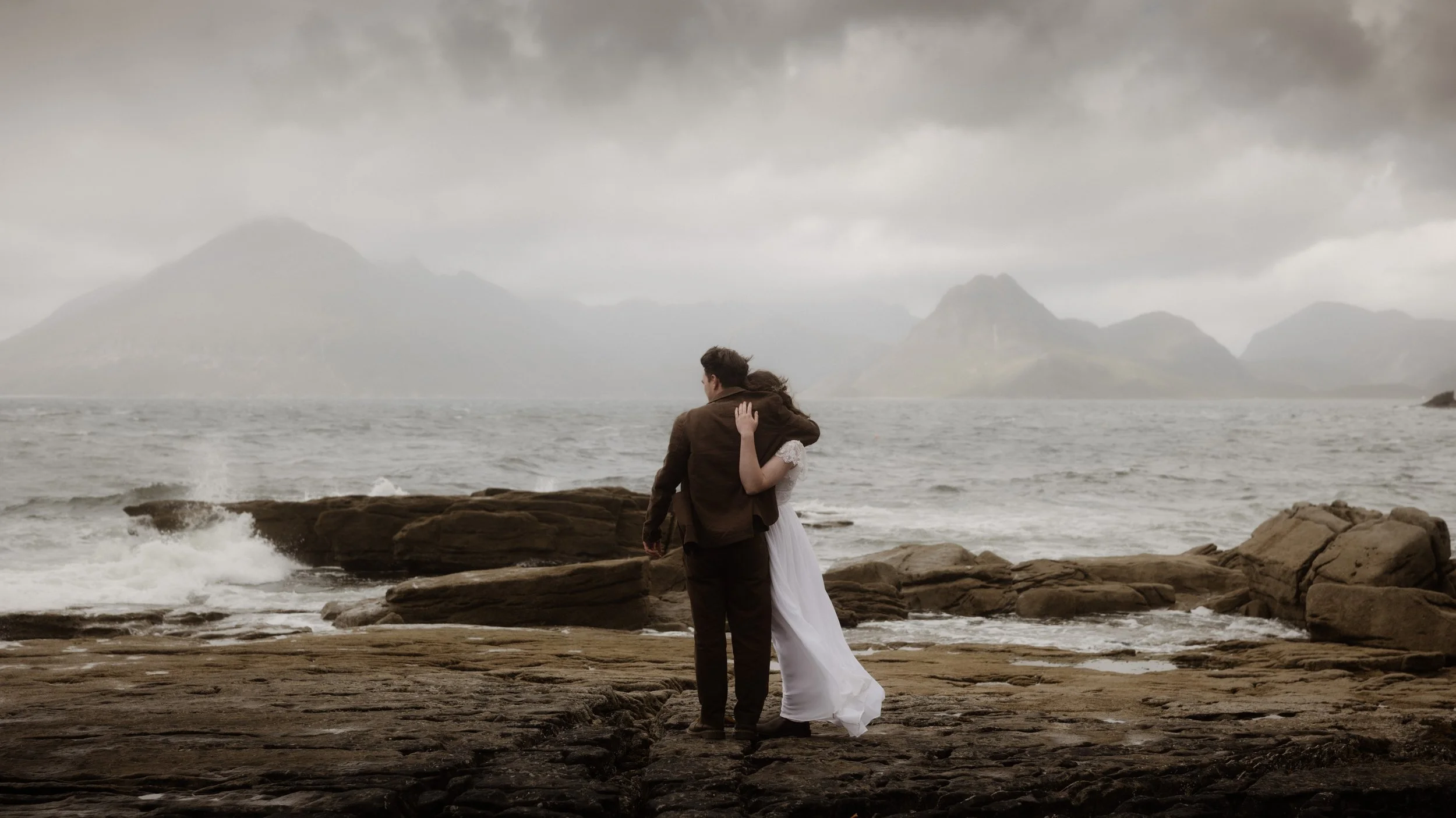 A couple dressed in wedding attire embrace on a rocky coastline under a cloudy sky, with mountains and the sea in the background.