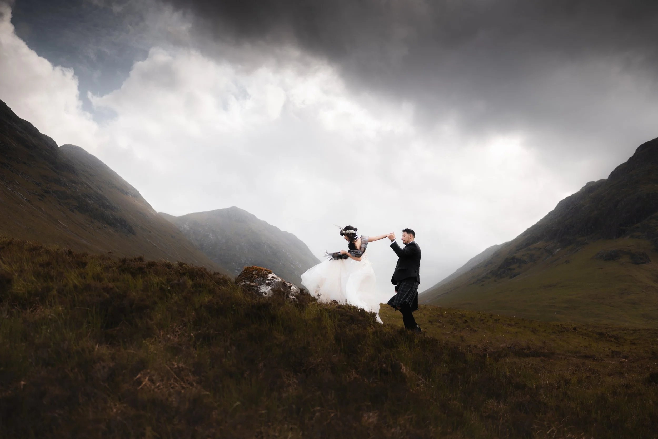 A couple dressed in wedding attire dancing on a grassy hillside in a mountainous landscape under a cloudy sky.