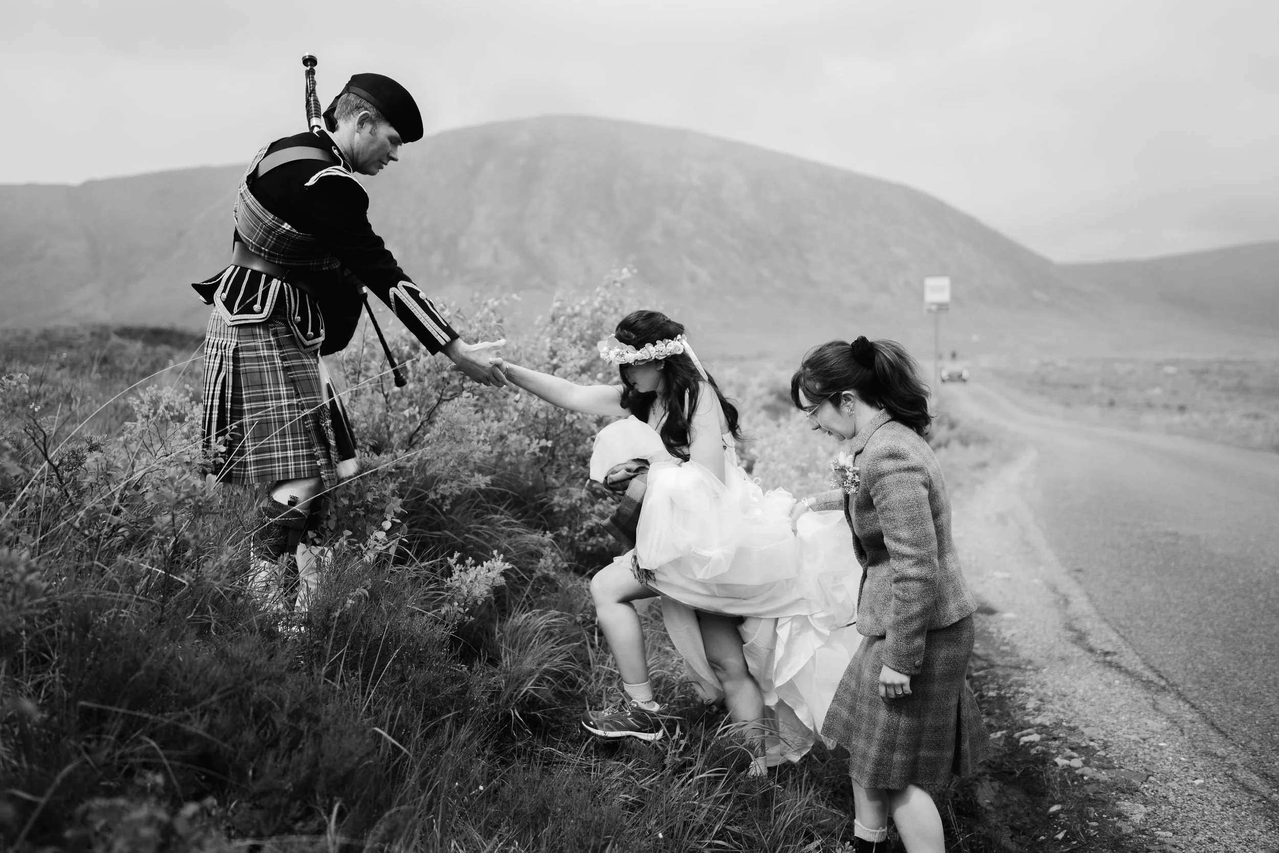 A black and white photo of a Scottish-themed wedding with a man in kilt helping a girl in a white dress, who is wearing a floral crown, onto a grassy hill. Another girl in a plaid skirt and blazer stands nearby, watching.