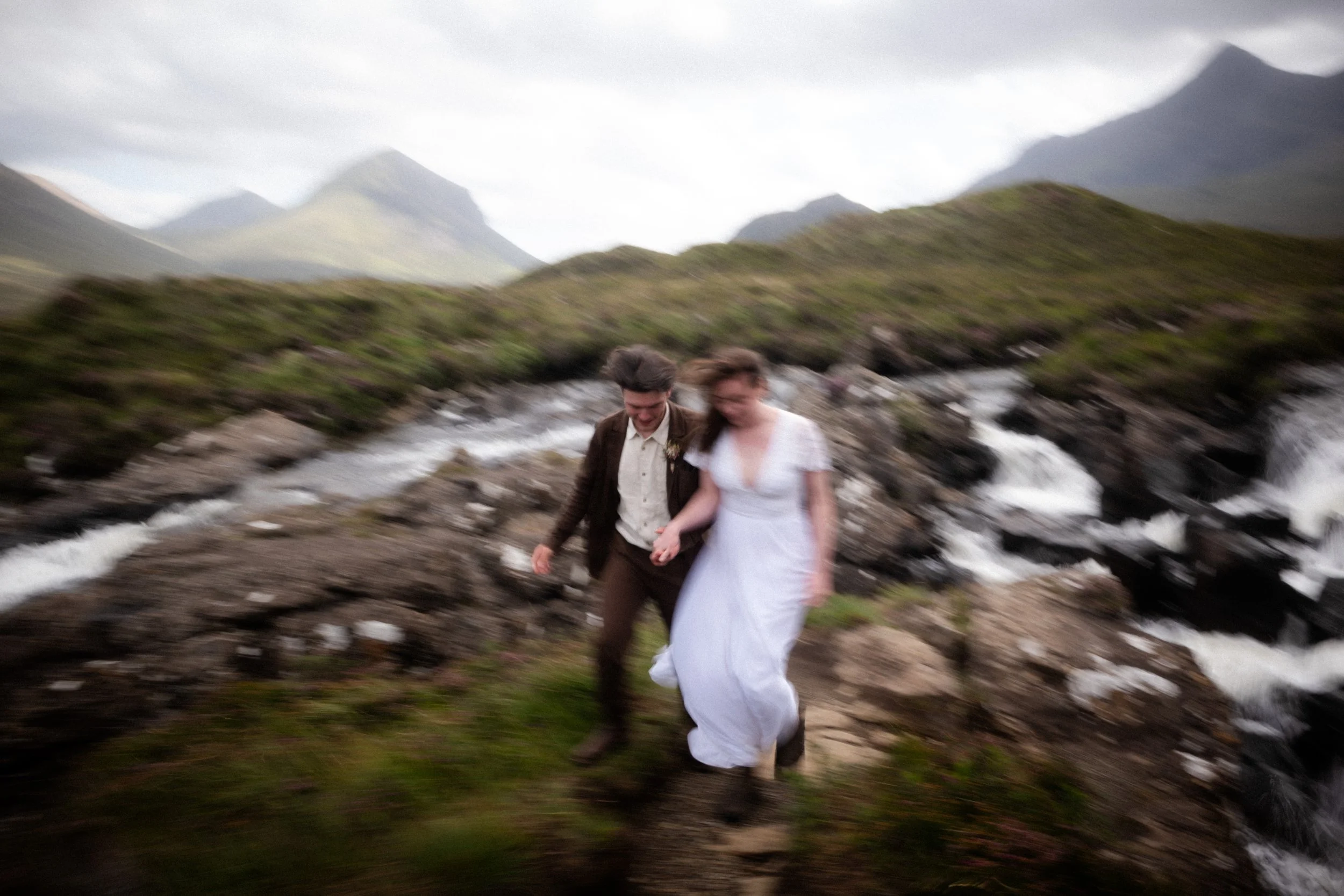 A blurred photo of a couple, possibly newlyweds, holding hands and walking beside a rushing river in a mountainous landscape with green hills and cloudy sky.