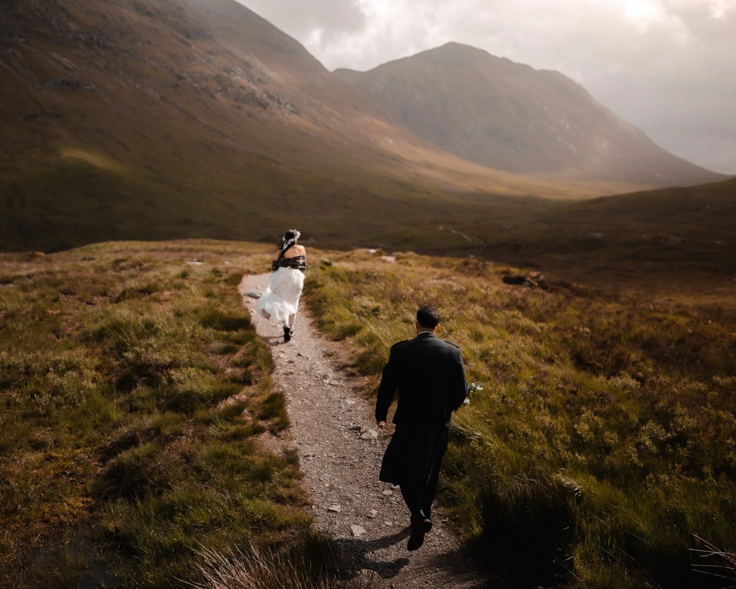 A couple, dressed in formal wedding attire, hiking on a dirt trail through a grassy valley surrounded by mountains with cloudy sky.