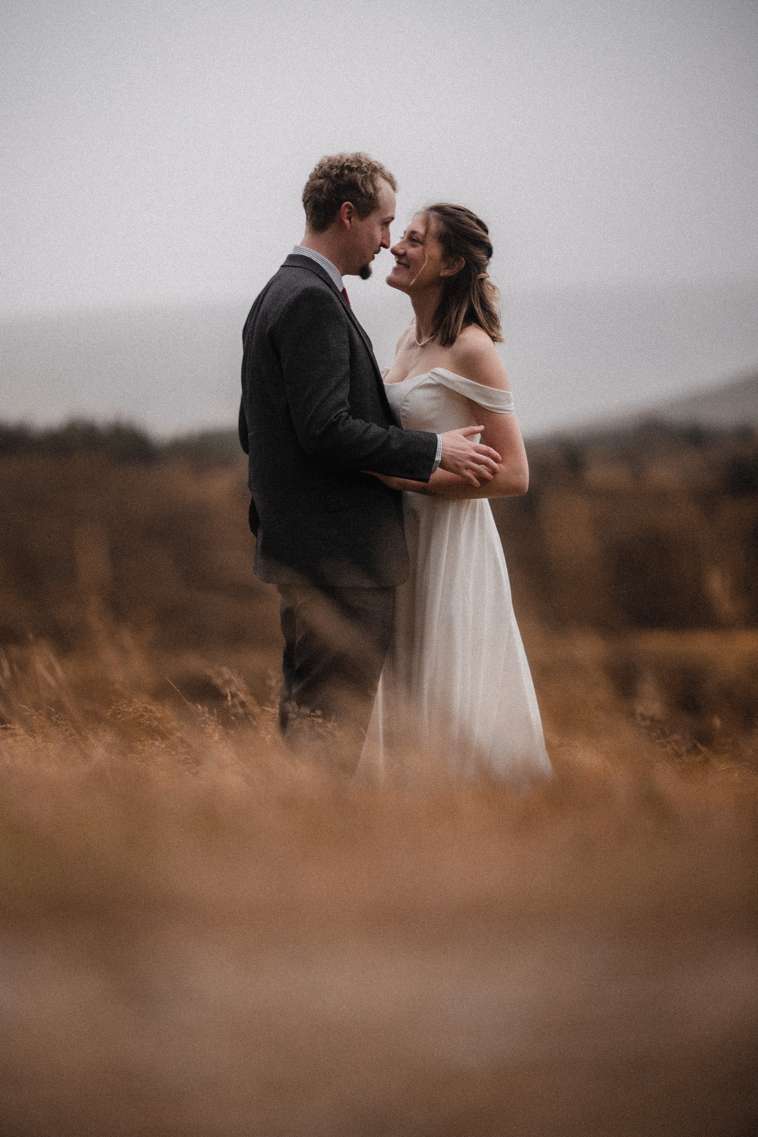 A couple in wedding attire standing in a grassy field, intimate and smiling at each other in Aviemore.