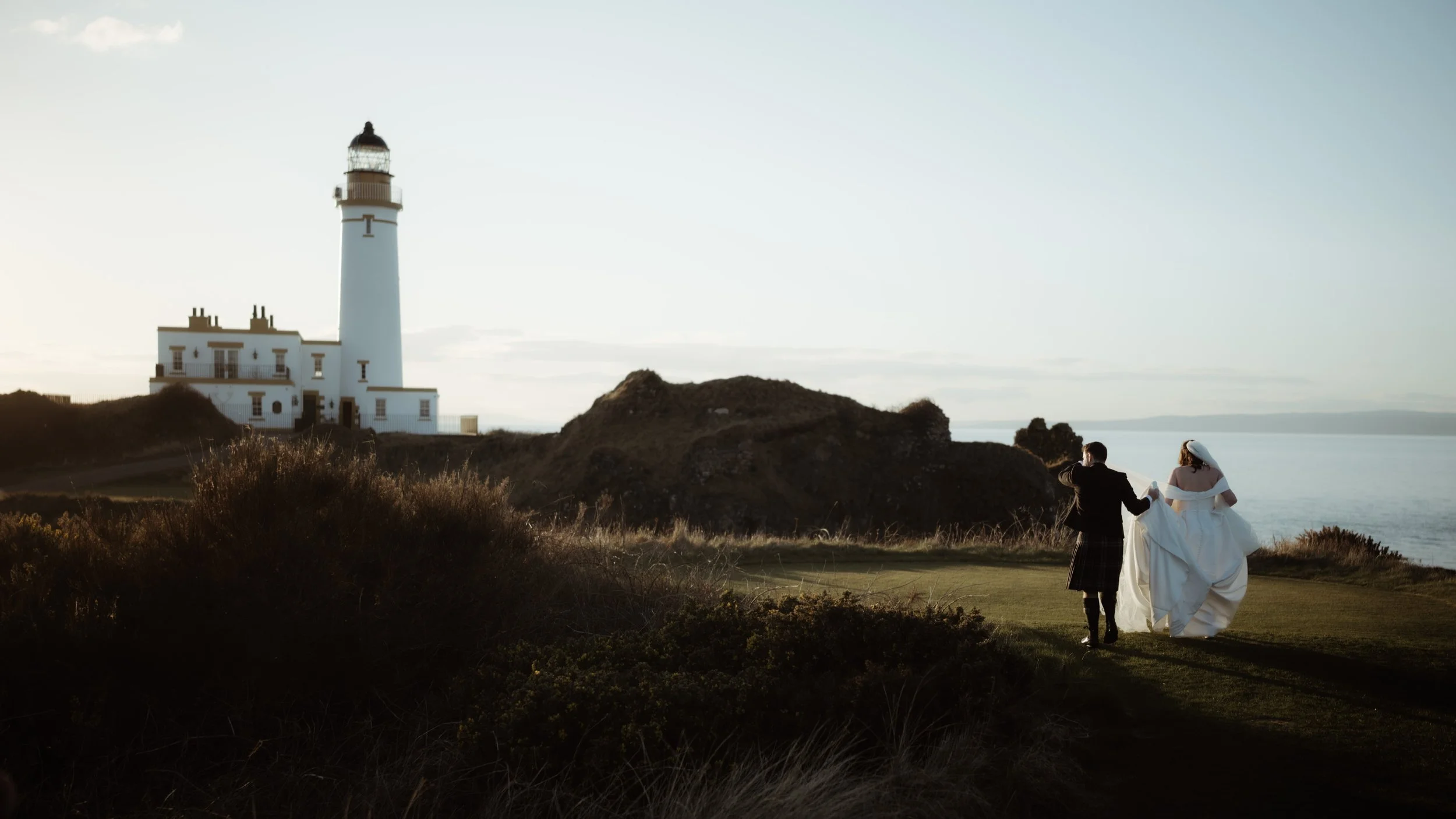 A bride and groom walking hand-in-hand near a lighthouse on a grassy coastal area as the sun sets.
