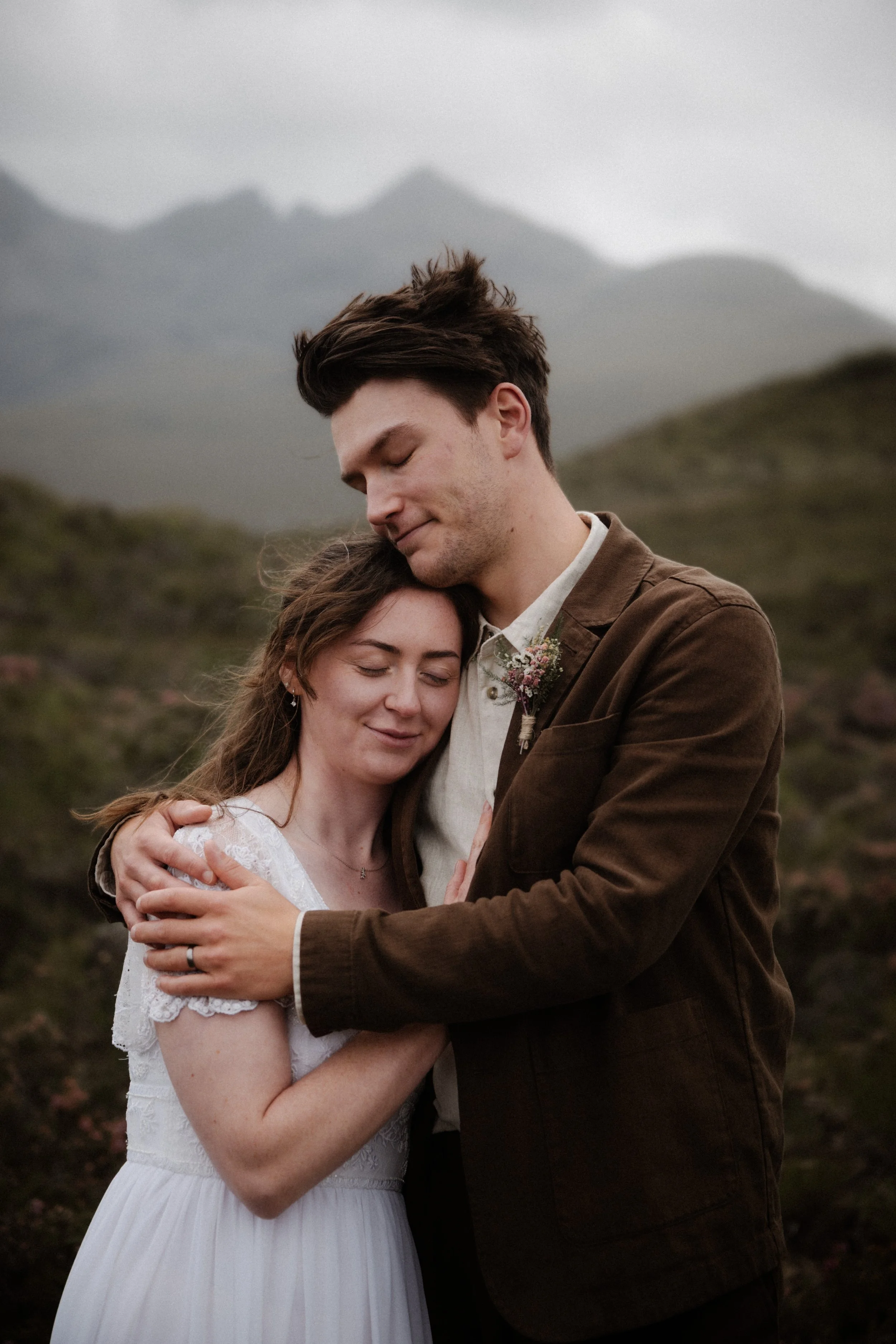 A couple embracing outdoors, with mountains in the background, during a wedding or romantic photo shoot.