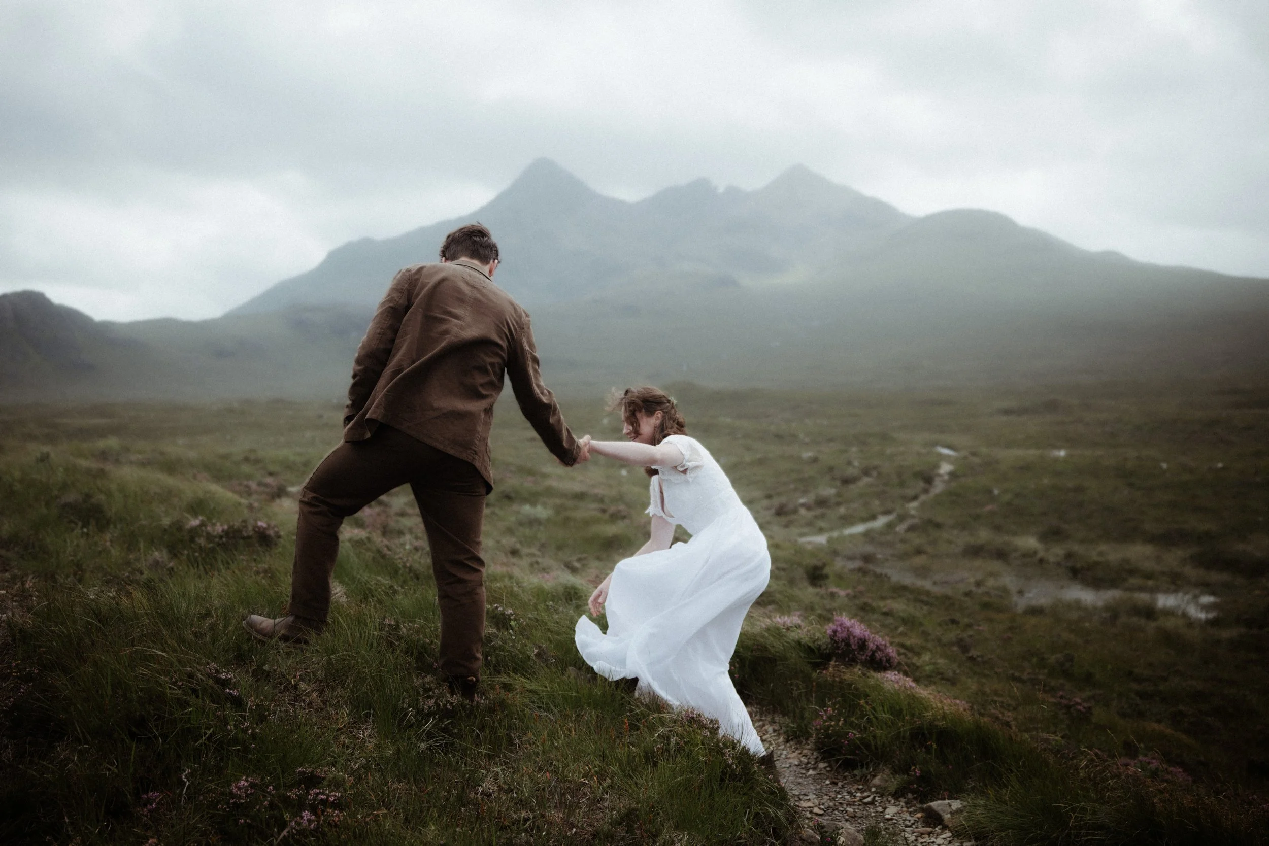A man and a woman holding hands in a grassy, mountainous landscape with overcast skies. The woman is wearing a white dress, and the man is in a brown jacket and pants.