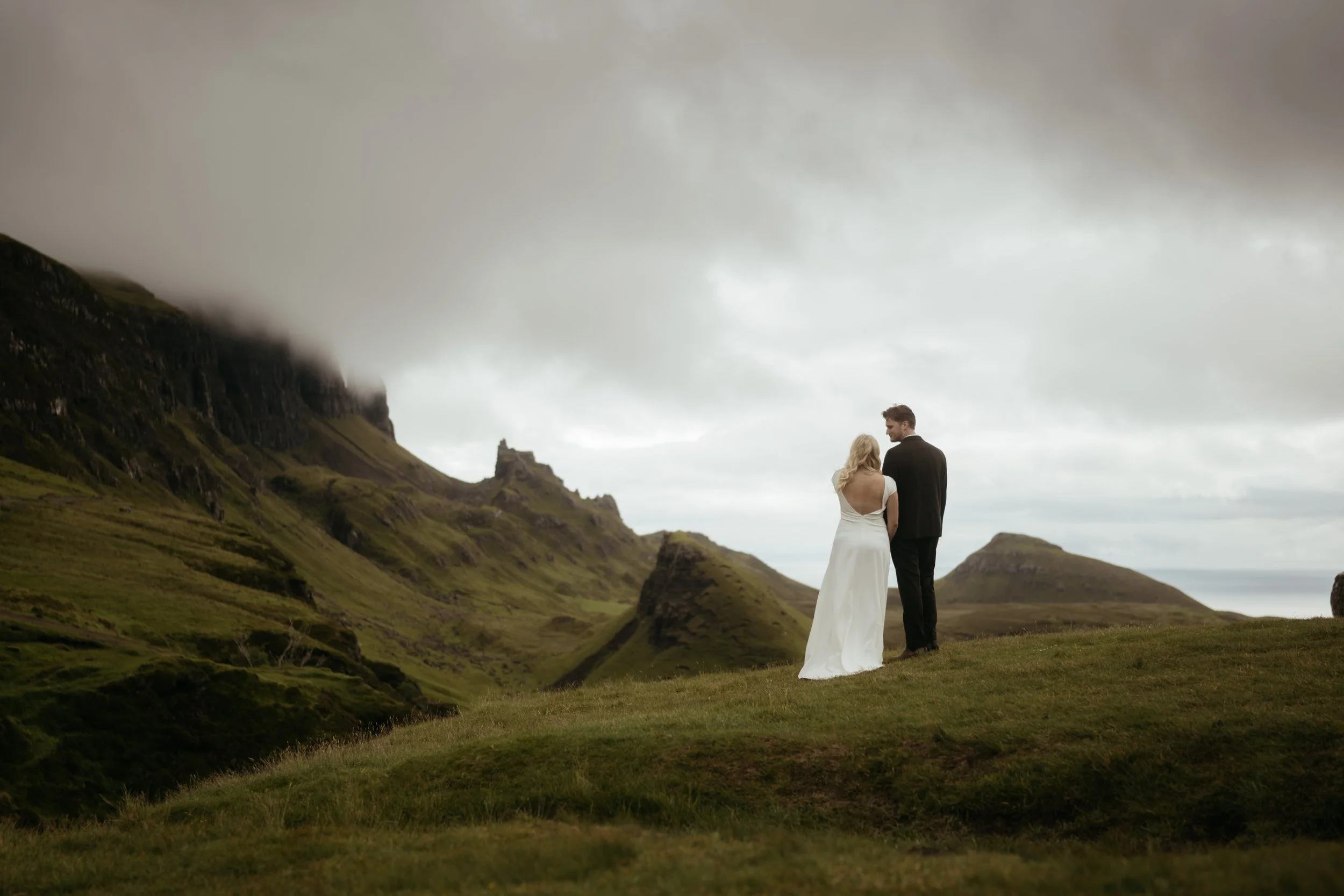 A bride and groom standing on a grassy hilltop overlooking a lush, green mountainous landscape under a cloudy sky.