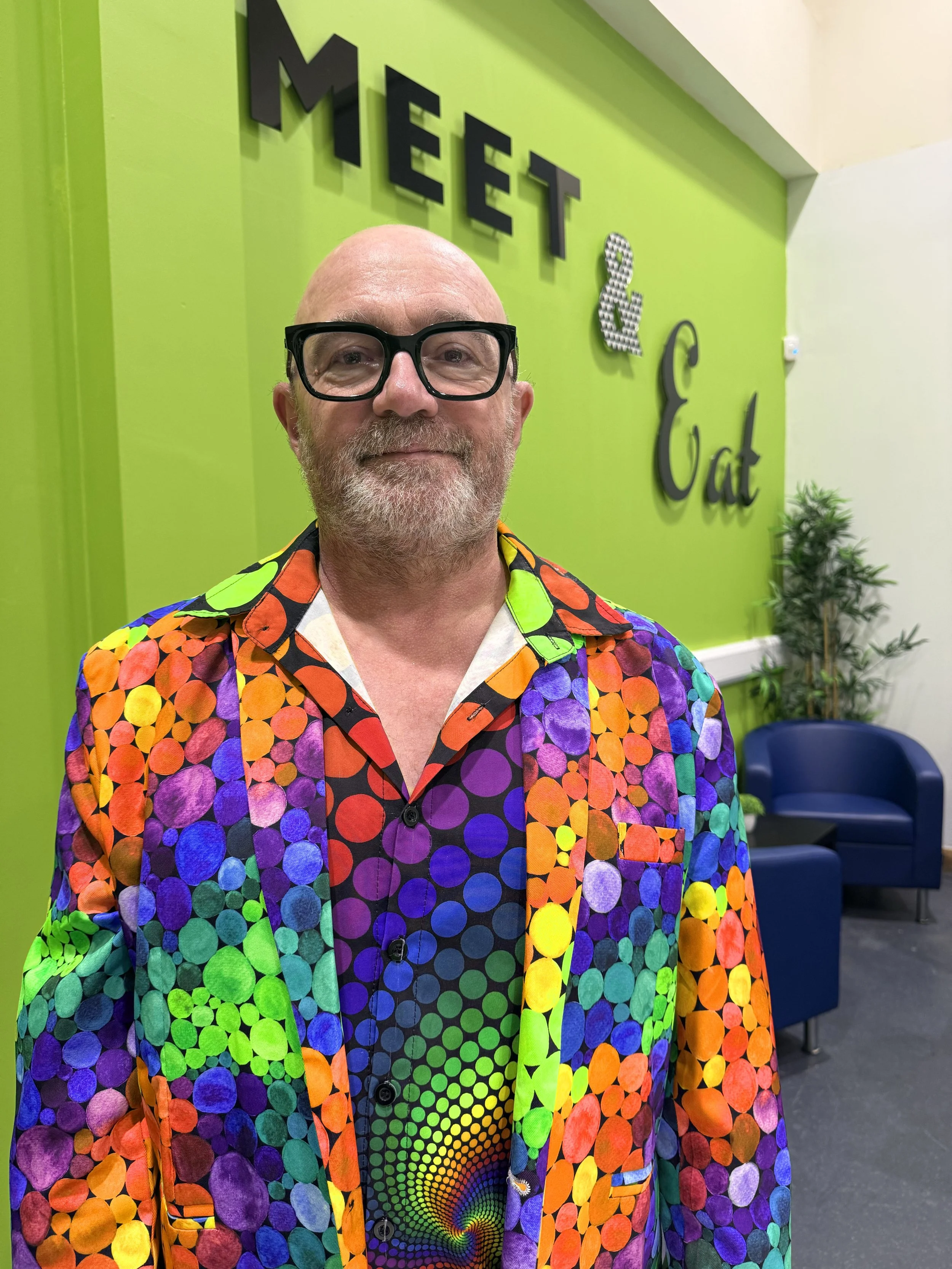 Stuart Lloyd Gould standing in front of a bright green wall with the words 'Meet & Eat' in decorative letters, wearing a colourful, patterned shirt.