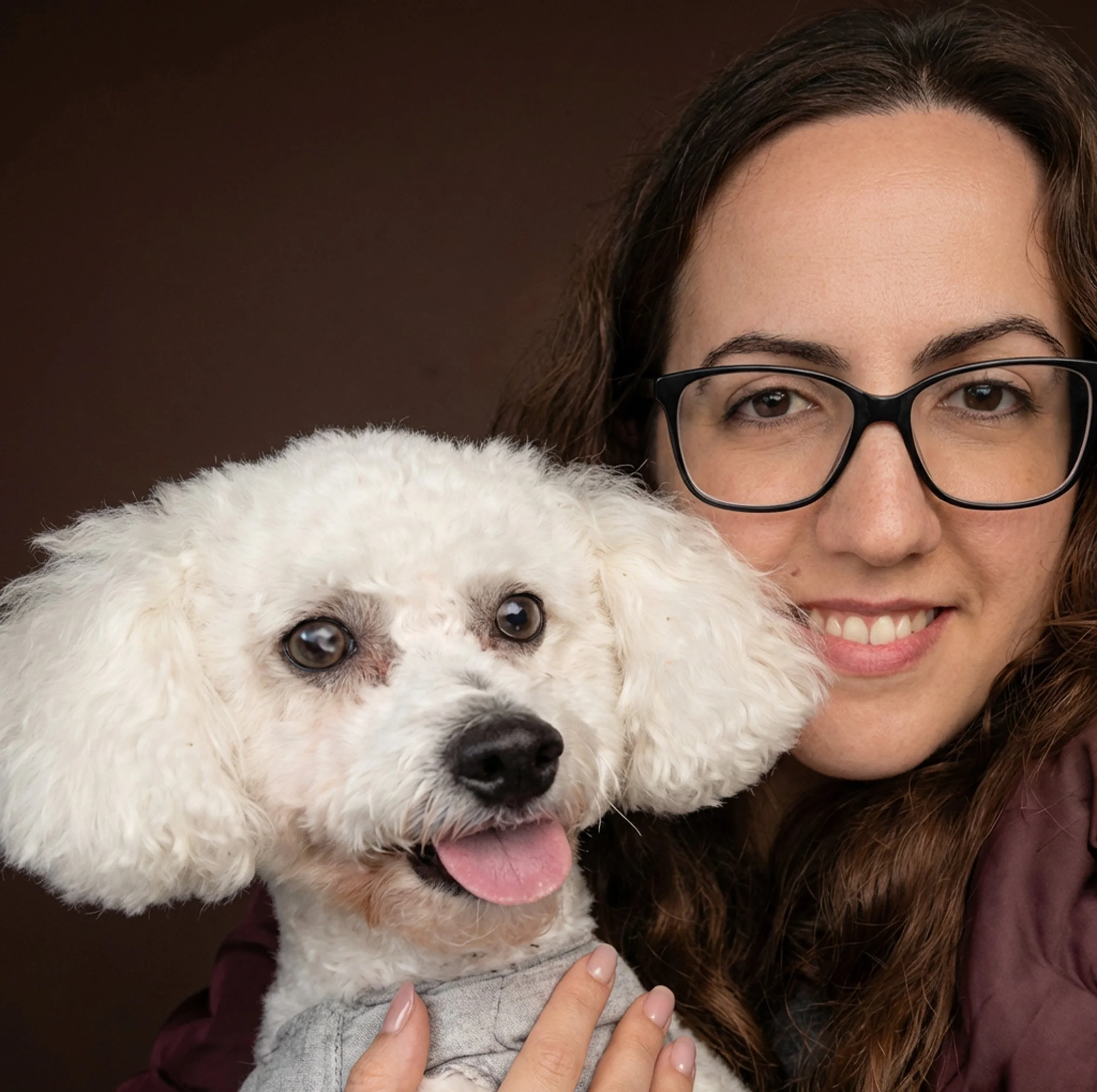 Marssa Peguinho smiling, holding a white poodle with fluffy hair, pink tongue out, and wearing a gray shirt, against a dark brown background.