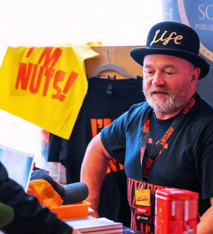 Stuart Lloyd Gould at a table with promotional materials, including a yellow sign reading 'I'M NUTS!'