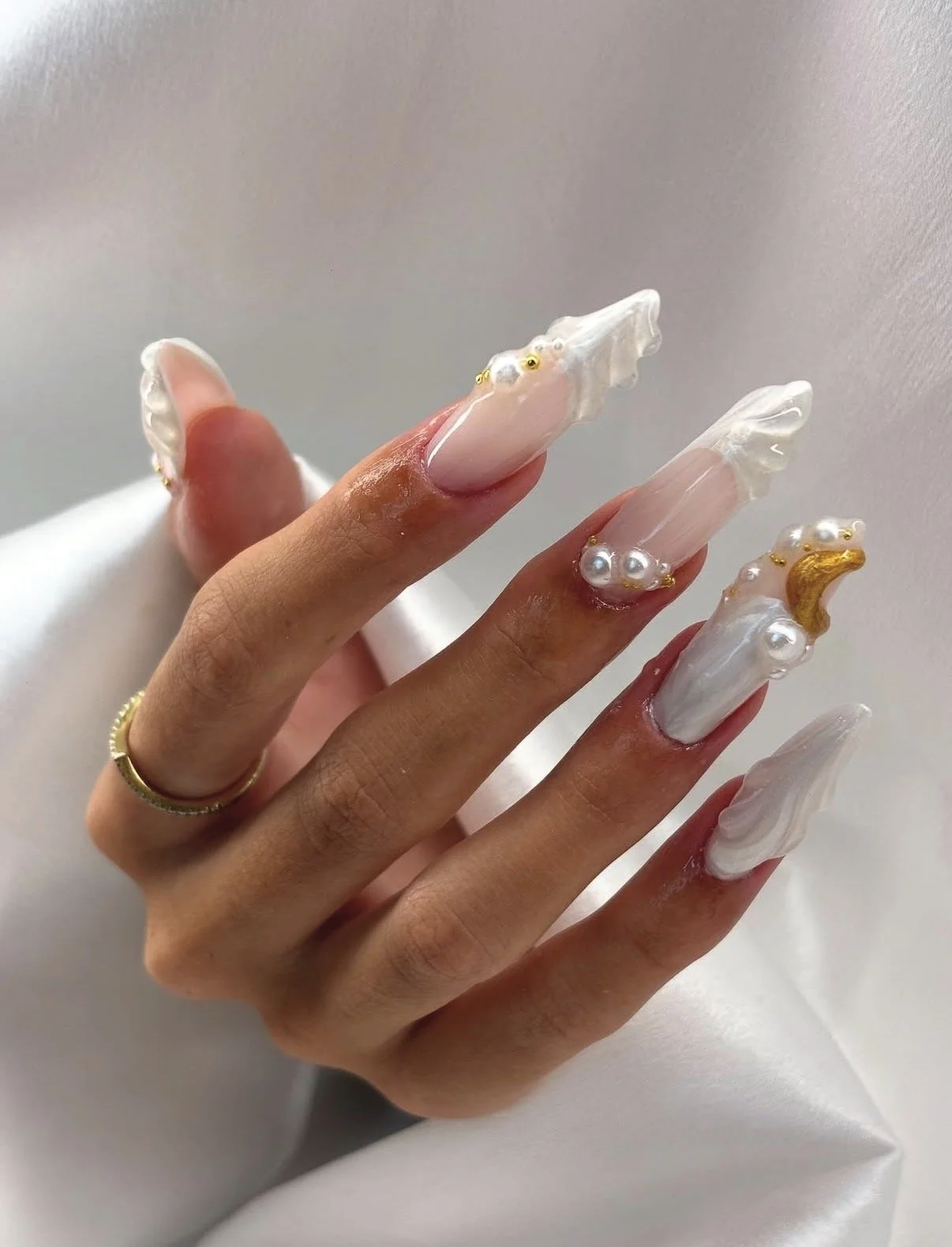 Close-up of a hand with long, decorated acrylic nails featuring white marble designs, pearl accents, and gold embellishments.