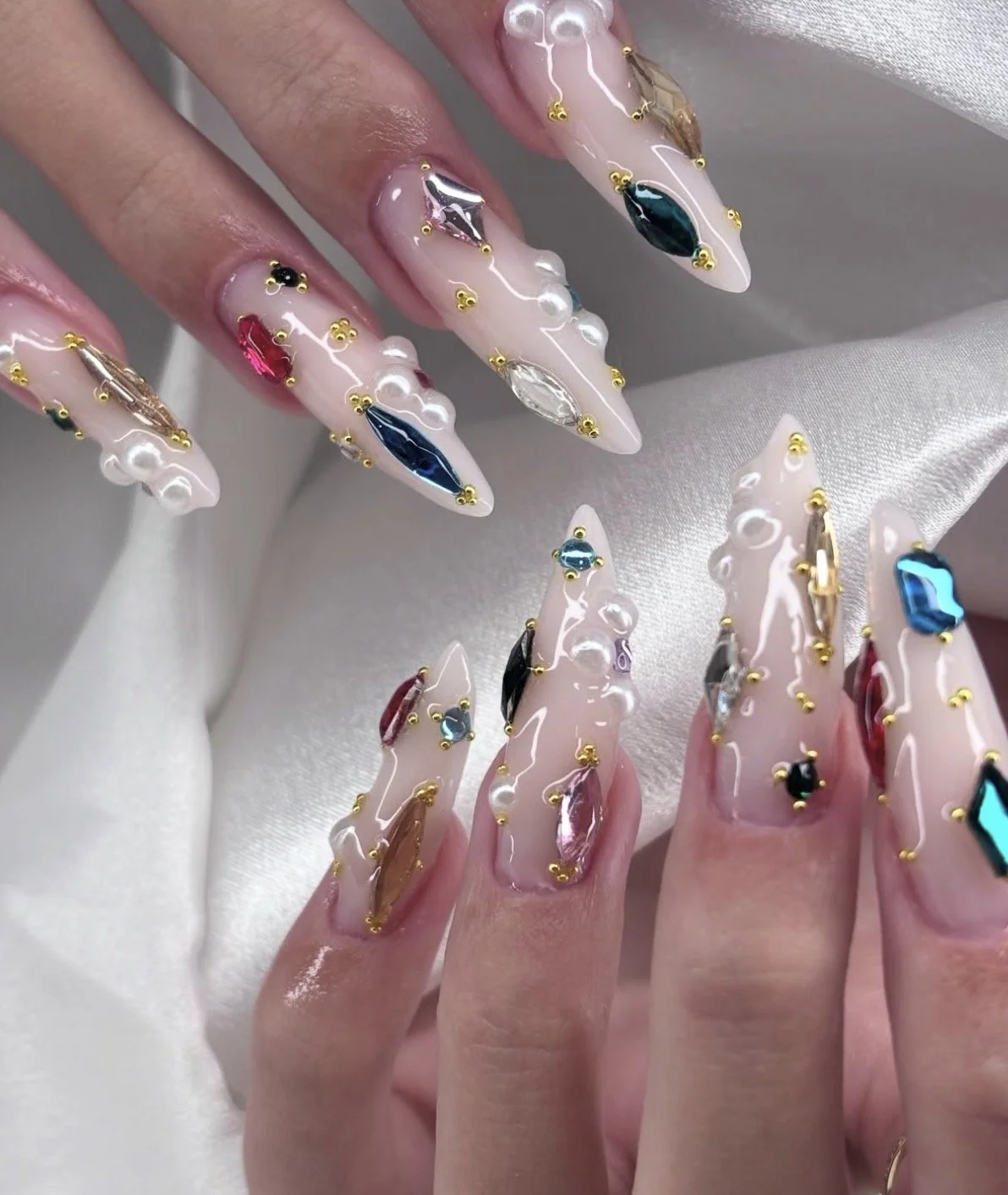 Close-up of long, pointed acrylic nails decorated with various colorful rhinestones and gold accents, against a soft, satin-like fabric background.