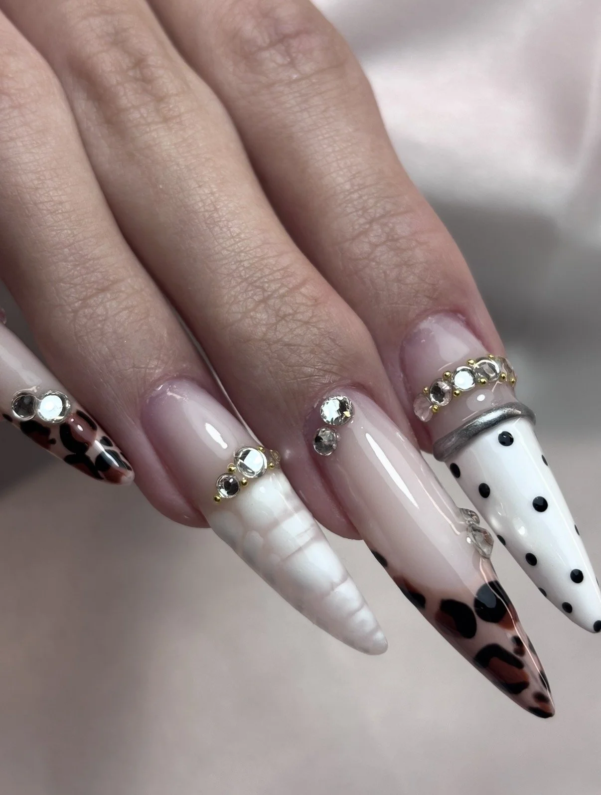 Close-up of a hand with long, pointed nails featuring various designs, rhinestones, and embellishments. The nails have animal print, pearls, and black polka dots with rhinestone accents.