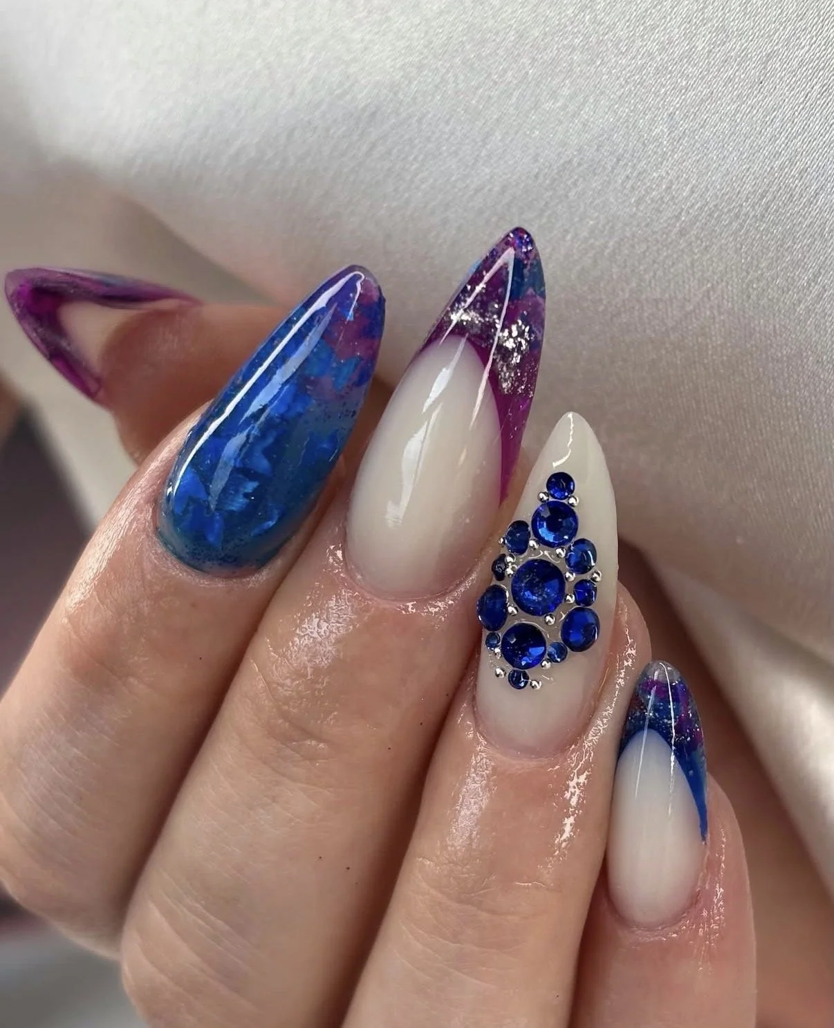 Close-up of a hand with elaborate gel nails featuring marbled purple and blue tips, a white and natural gradient design, and a nail adorned with blue rhinestones.