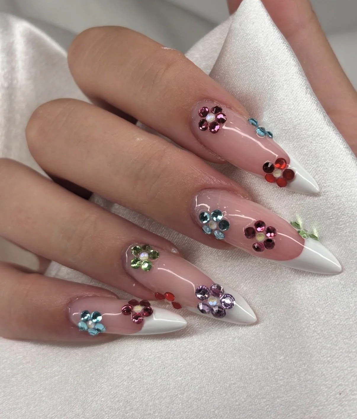 Close-up of a hand with long, pointed, acrylic nails decorated with multicolored rhinestones arranged in flower shapes on a pink base with white tips.
