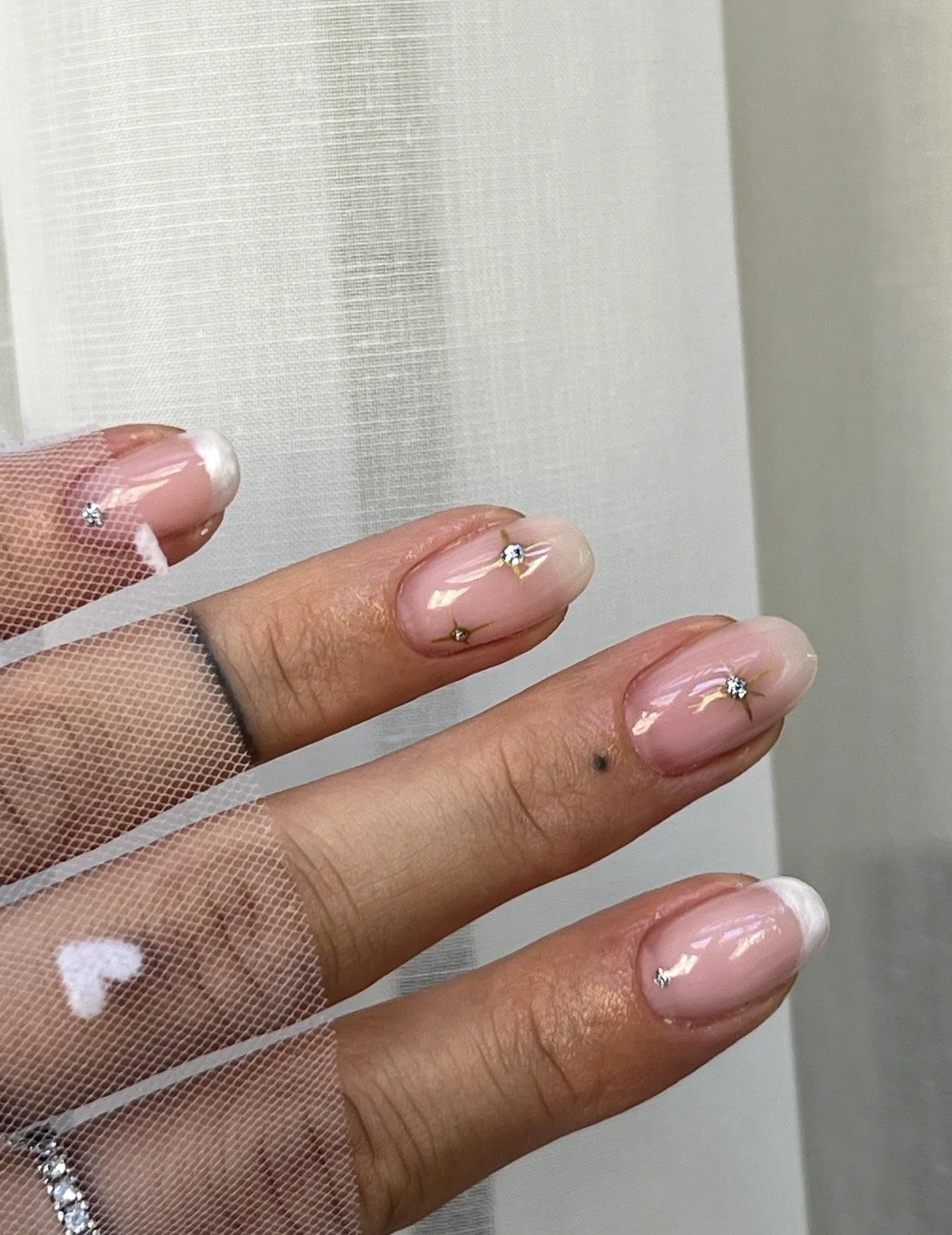 Close-up of a hand with decorated nails, each featuring a small rhinestone, against a white background, with one finger slightly raised.