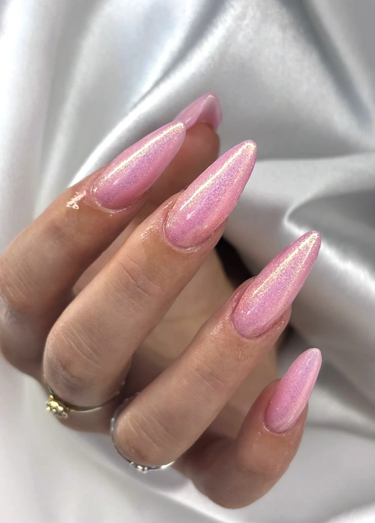 Close-up of a hand with long, pink, iridescent, almond-shaped nails, resting on a white surface with a satin fabric background.