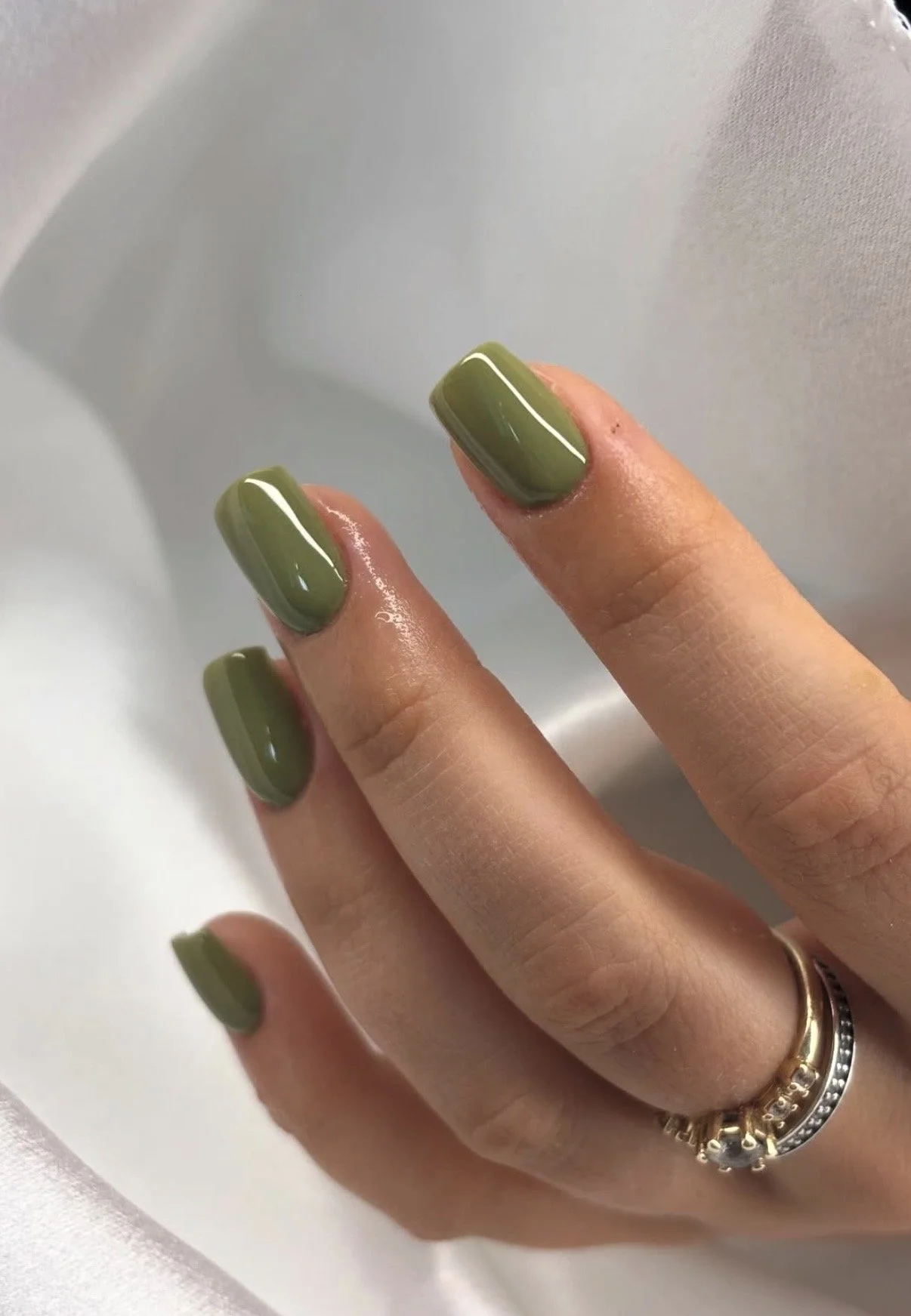 Close-up of a hand with olive green painted nails, wearing two rings on the ring finger, against a light-colored background.