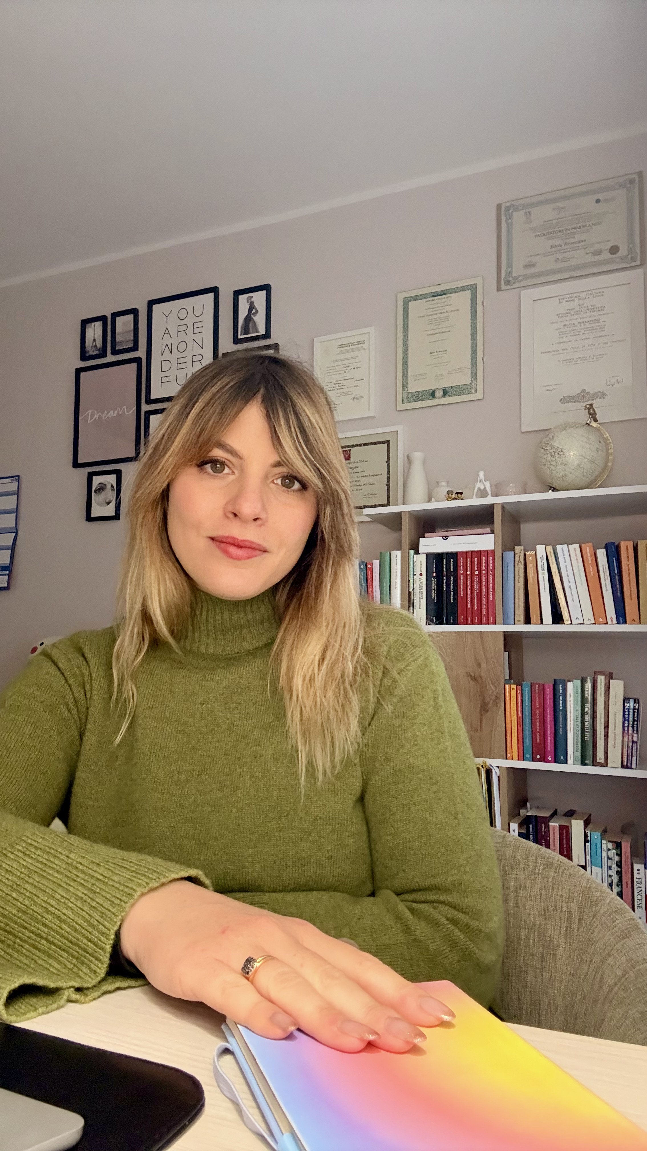 Una donna con capelli castani e vestito verde seduta ad un tavolo in una stanza con scaffali pieni di libri e quadri alle pareti.