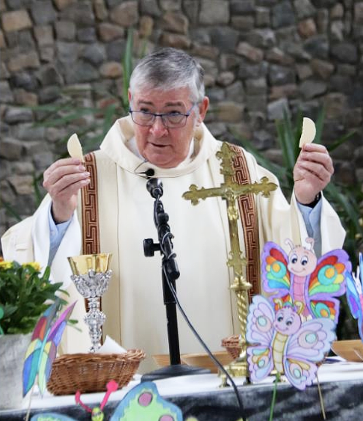 A priest dressed in white robes stands behind a table decorated with butterflies and flowers, holding a cracker in each hand during a religious ceremony, with a stone wall in the background.