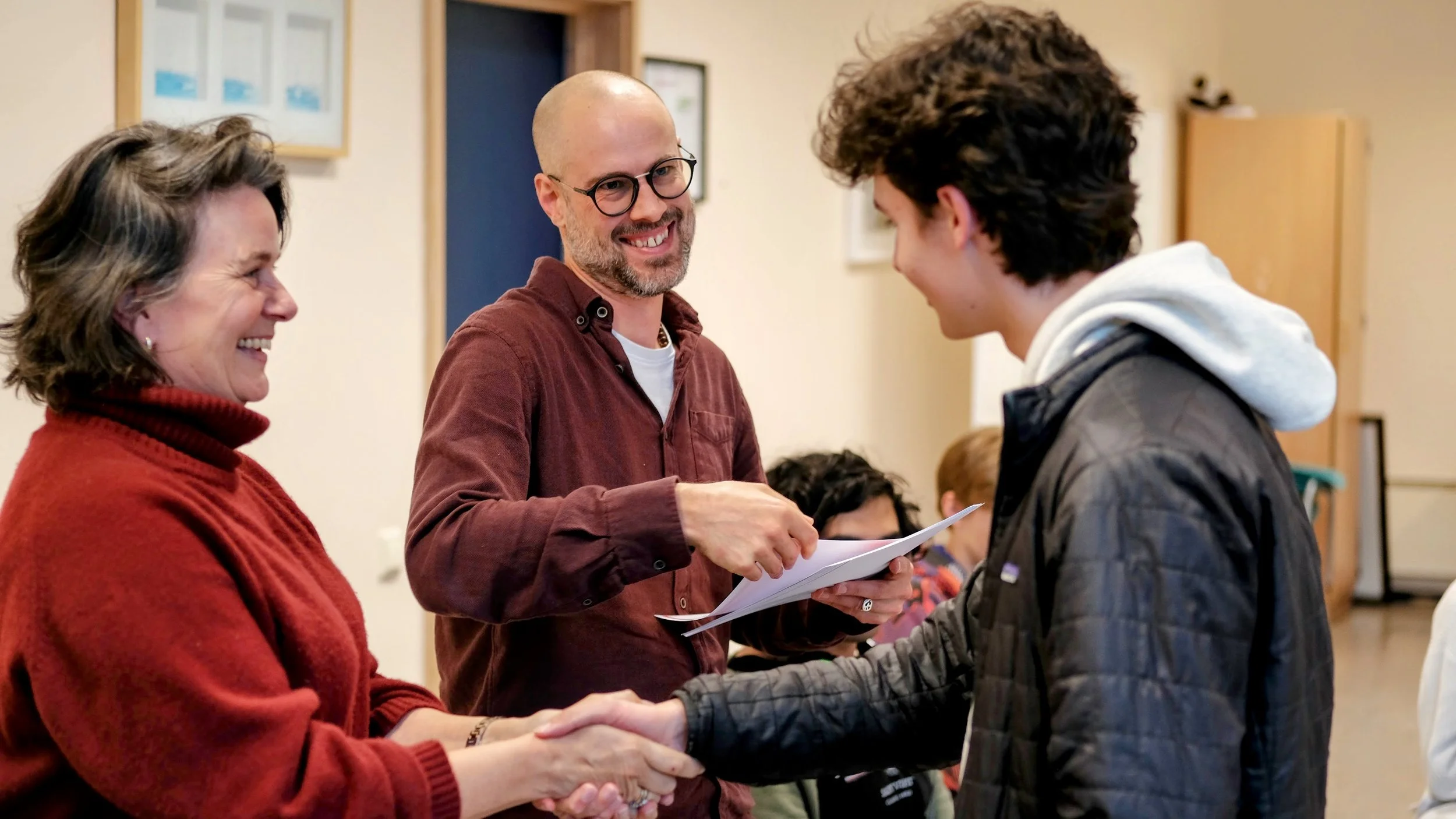 A woman and a man shaking hands at a formal indoor event, with another man holding a certificate in the background. All three are smiling.