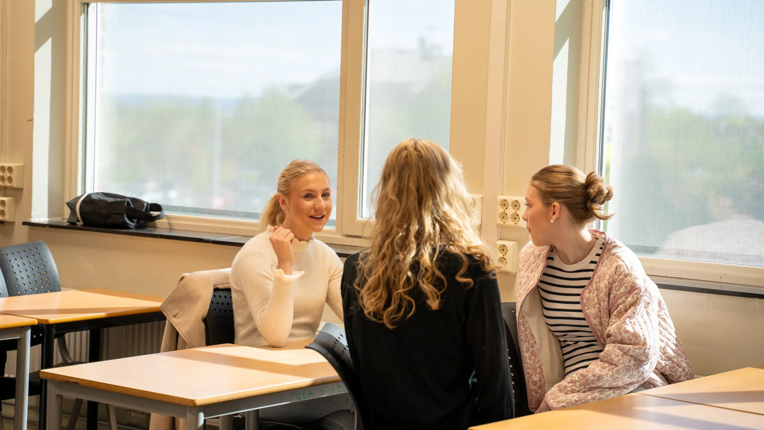 Three young women sitting at desks in a classroom, engaged in conversation near large windows.