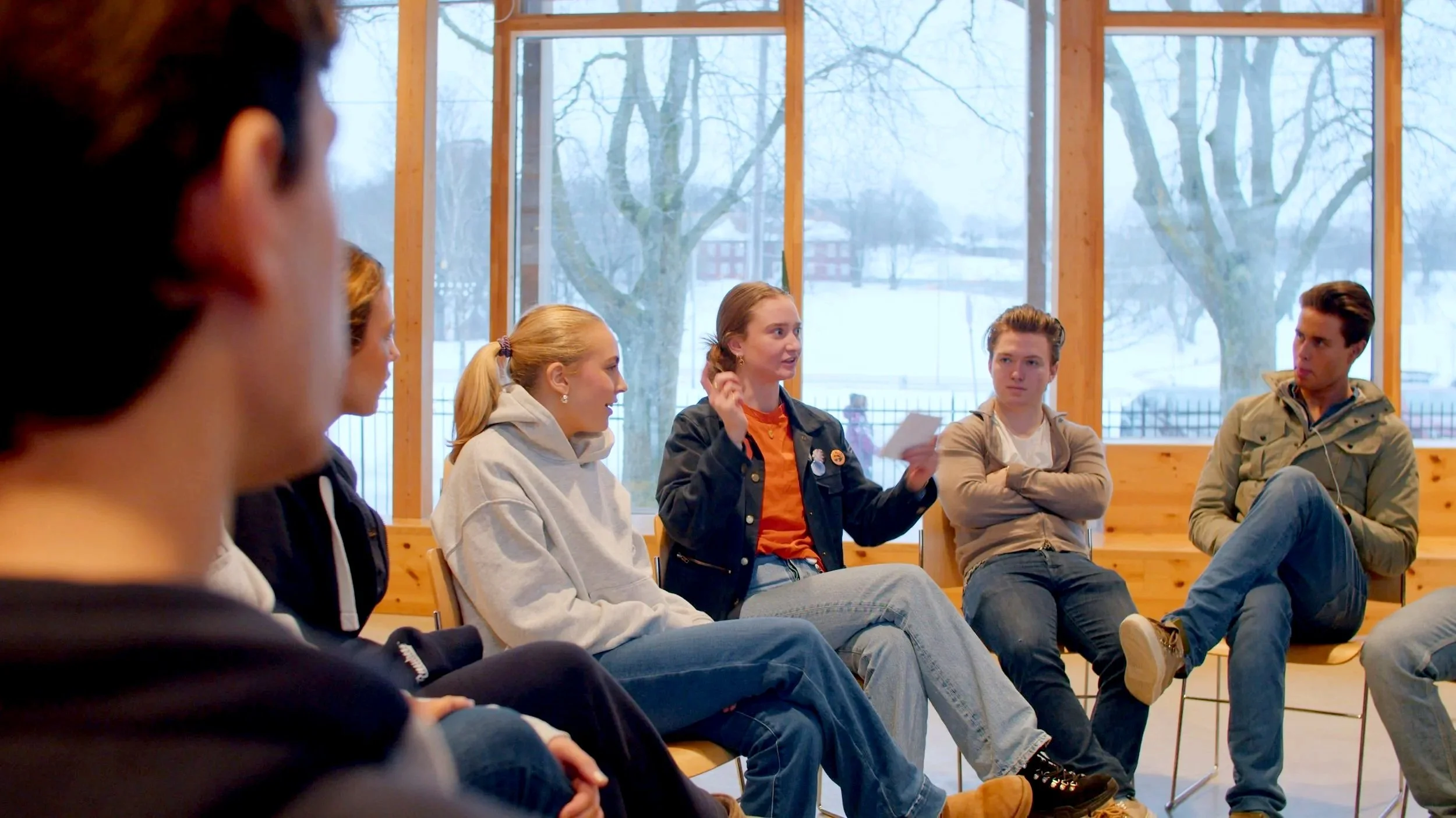 Group of young people sitting in a circle, engaged in conversation indoors, with large windows showing snowy trees outside.