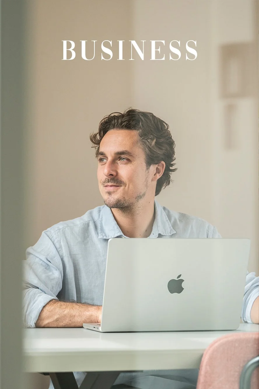 Man zit aan een tafel met een open MacBook op schoot, kijkt nadenkend in de verte, met de tekst 'BUSINESS' erboven.
